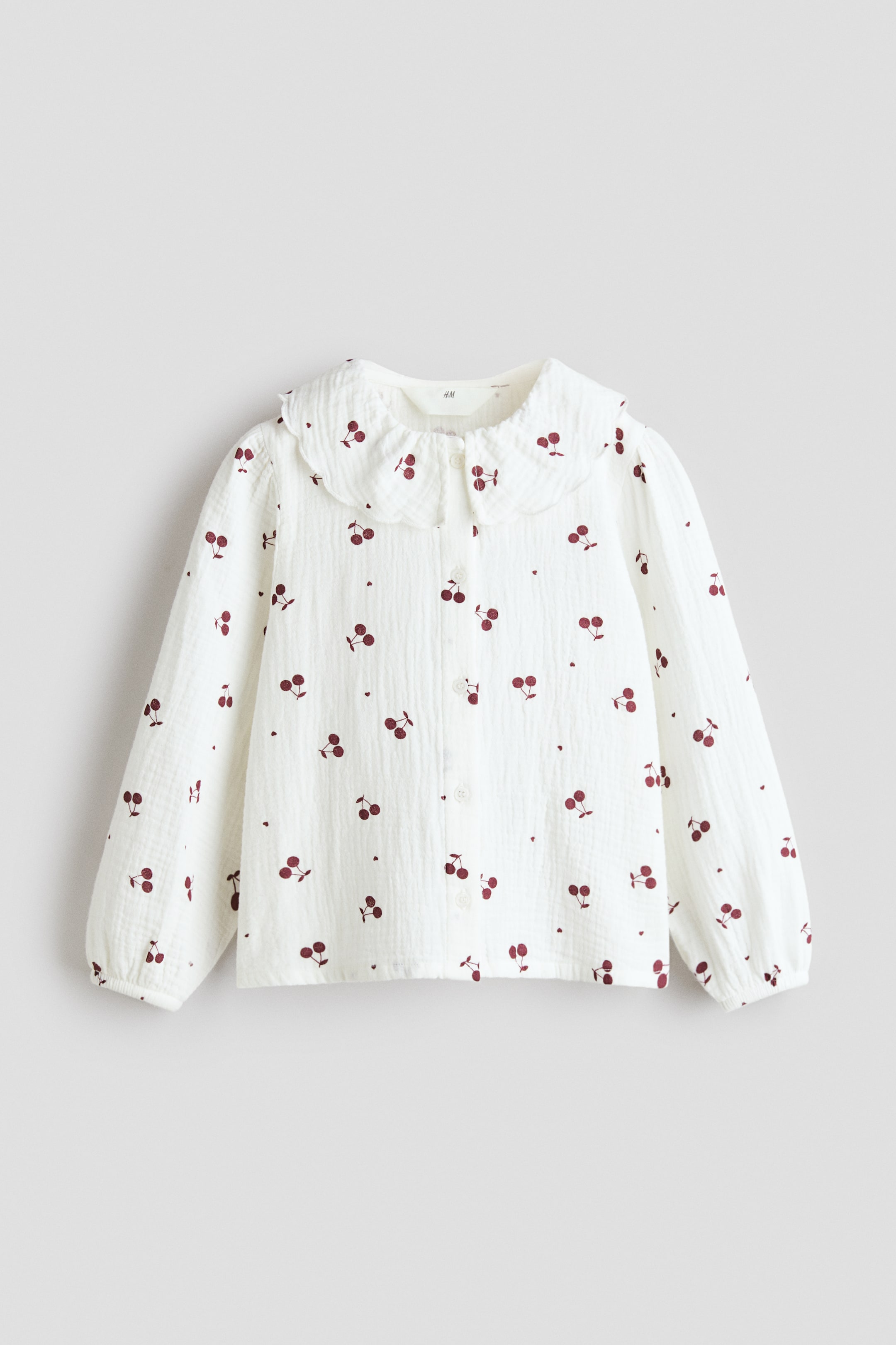 View larger image: A white textured blouse showcases a red cherry pattern, complemented by a scalloped Peter Pan collar and a full-length button-front closure. Crafted with long sleeves that conclude in elasticated cuffs, this piece is both stylish and functional.