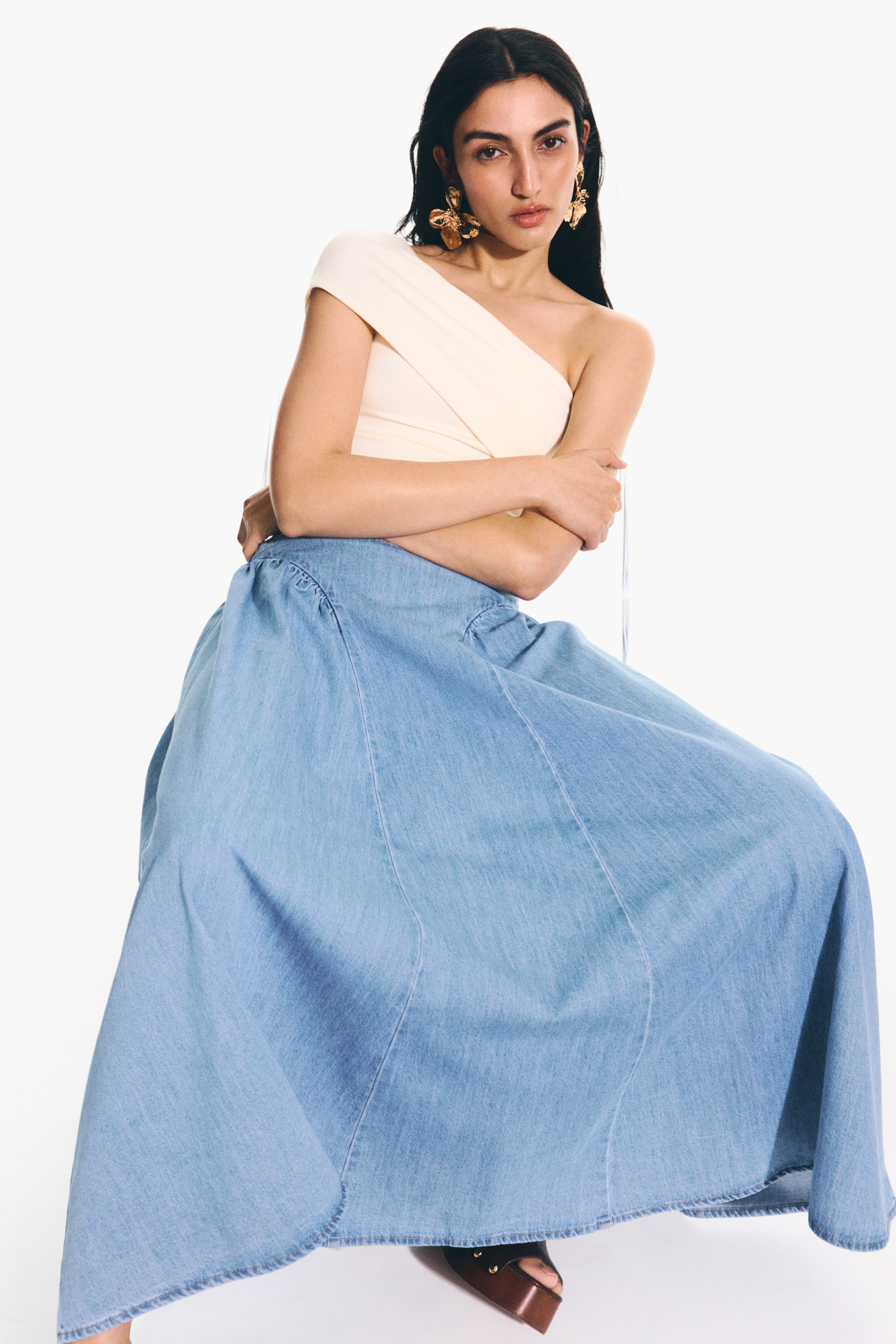 Ampliar la imagen: Una falda maxi de mezclilla azul claro presenta una silueta A-line completa y fluida con costuras verticales visibles en los paneles. Muestra una cintura fruncida, creando un drapeado voluminoso a medida que se extiende hacia el suelo.