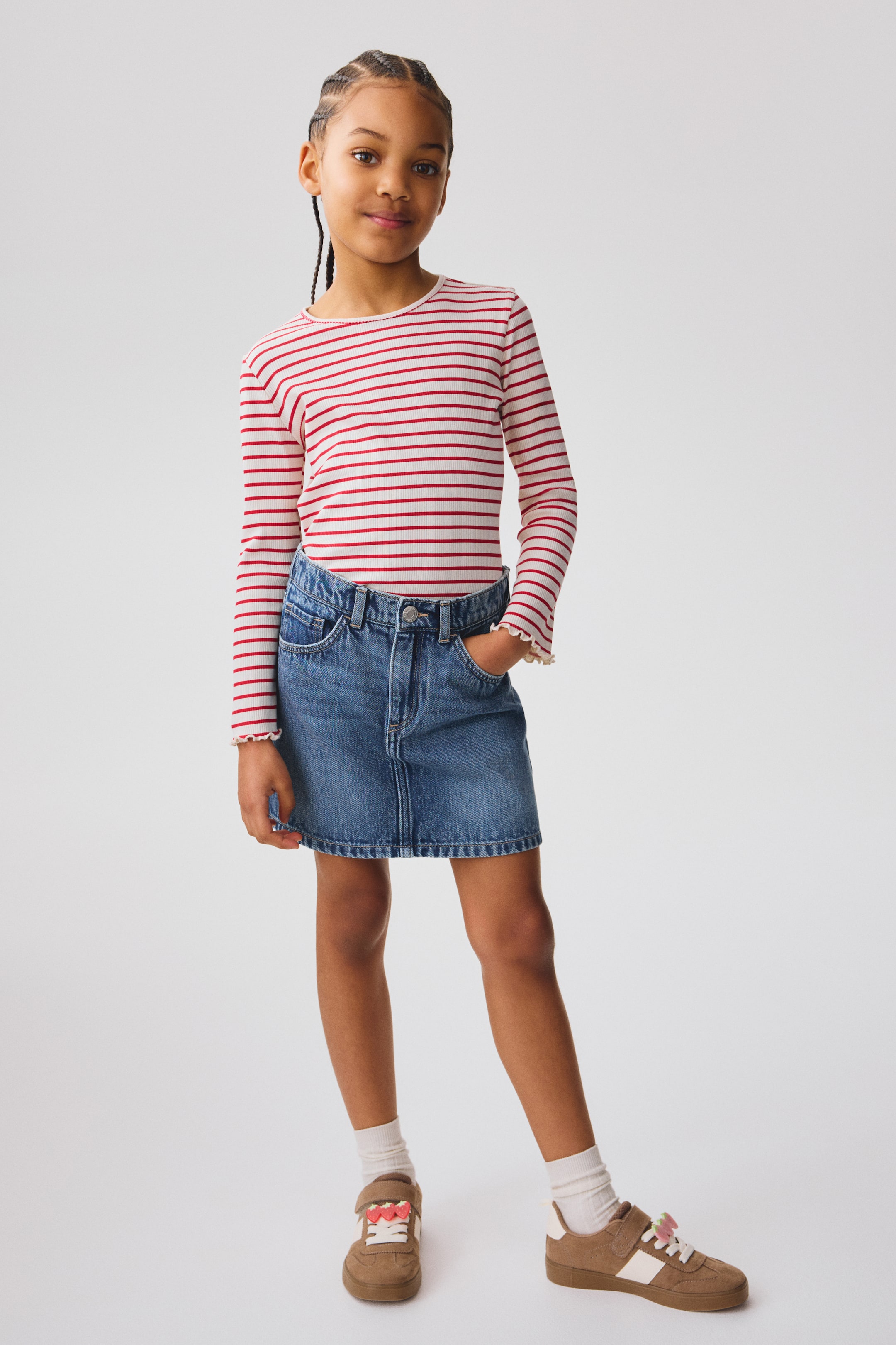 הצגת תמונה גדולה יותר: חולצת ילדים עם שרוולים ארוכים ומרקם מצולע מציגה פסים אופקיים בצבעים אדום ולבן, צווארון עם קפלים ושרוולים עם קפלים, והיא נלבשת בתוך חצאית מיני ג'ינס כחולה עם כיסים קדמיים וסגירת כפתורים גלויה במותן.