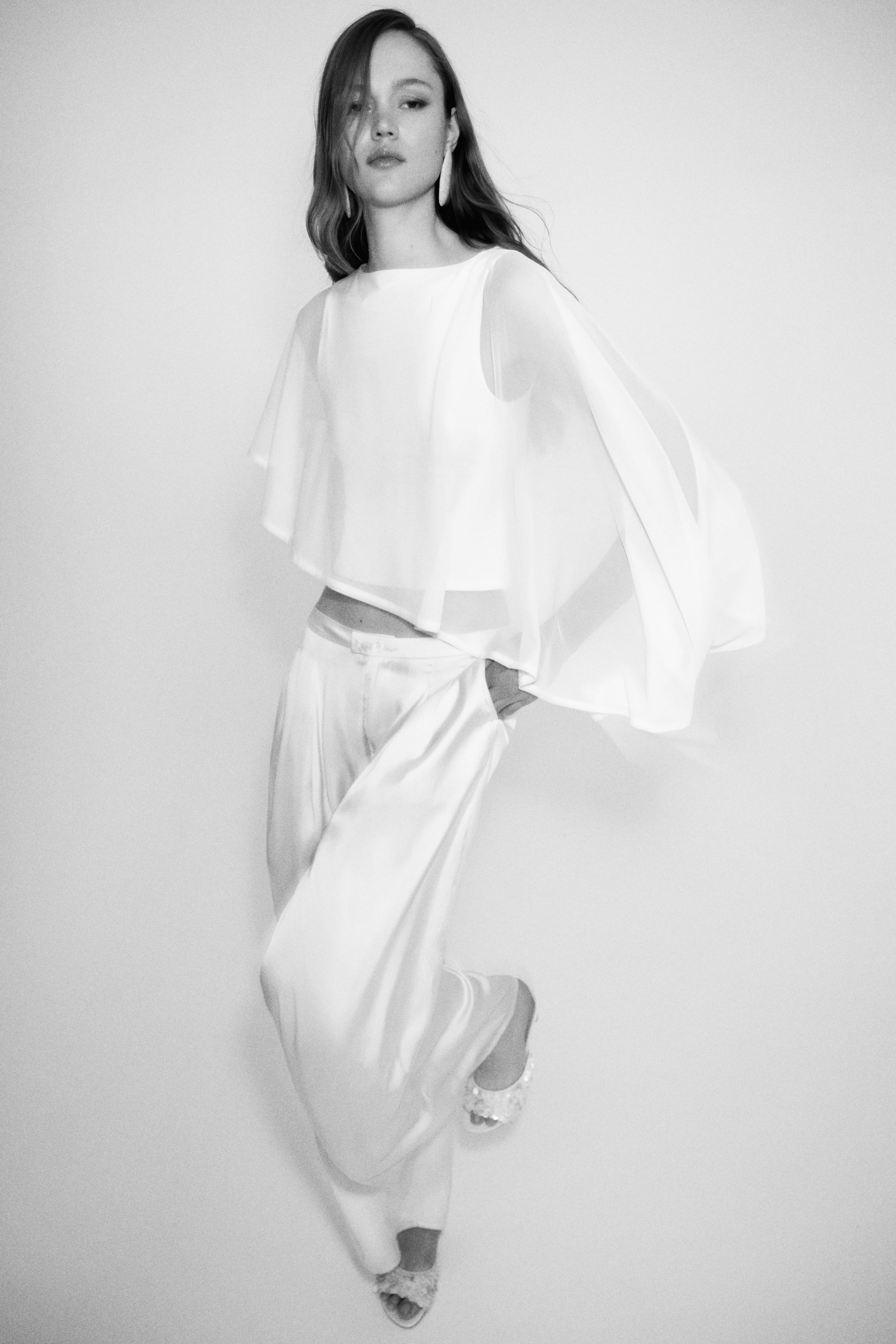 View larger image: A white sleeveless top featuring a sheer, flowing cape-like overlay is showcased alongside matching high-waisted, wide-leg pants. The lustrous fabric of the pants drapes fluidly, with one hand resting in a side pocket, viewed from a slightly angled front.