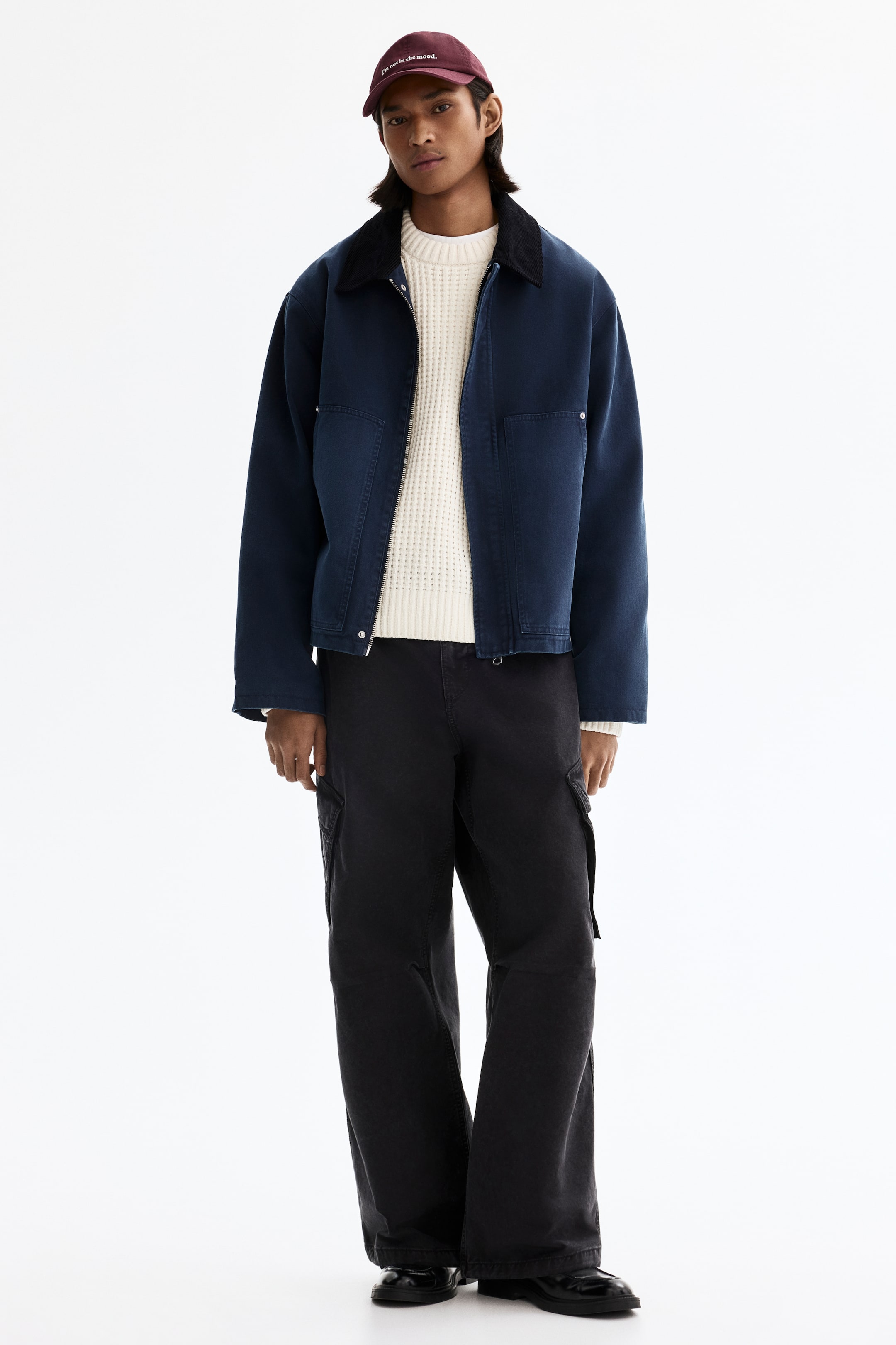 View larger image: A full outfit is being presented, featuring a maroon baseball cap with white embroidered text and a dark blue workwear jacket with a corduroy collar. The jacket is layered over a cream waffle-knit crewneck sweater, with black wide-leg cargo pants showing visible side pockets and black polished loafers completing the ensemble. The outfit is shown facing forward.