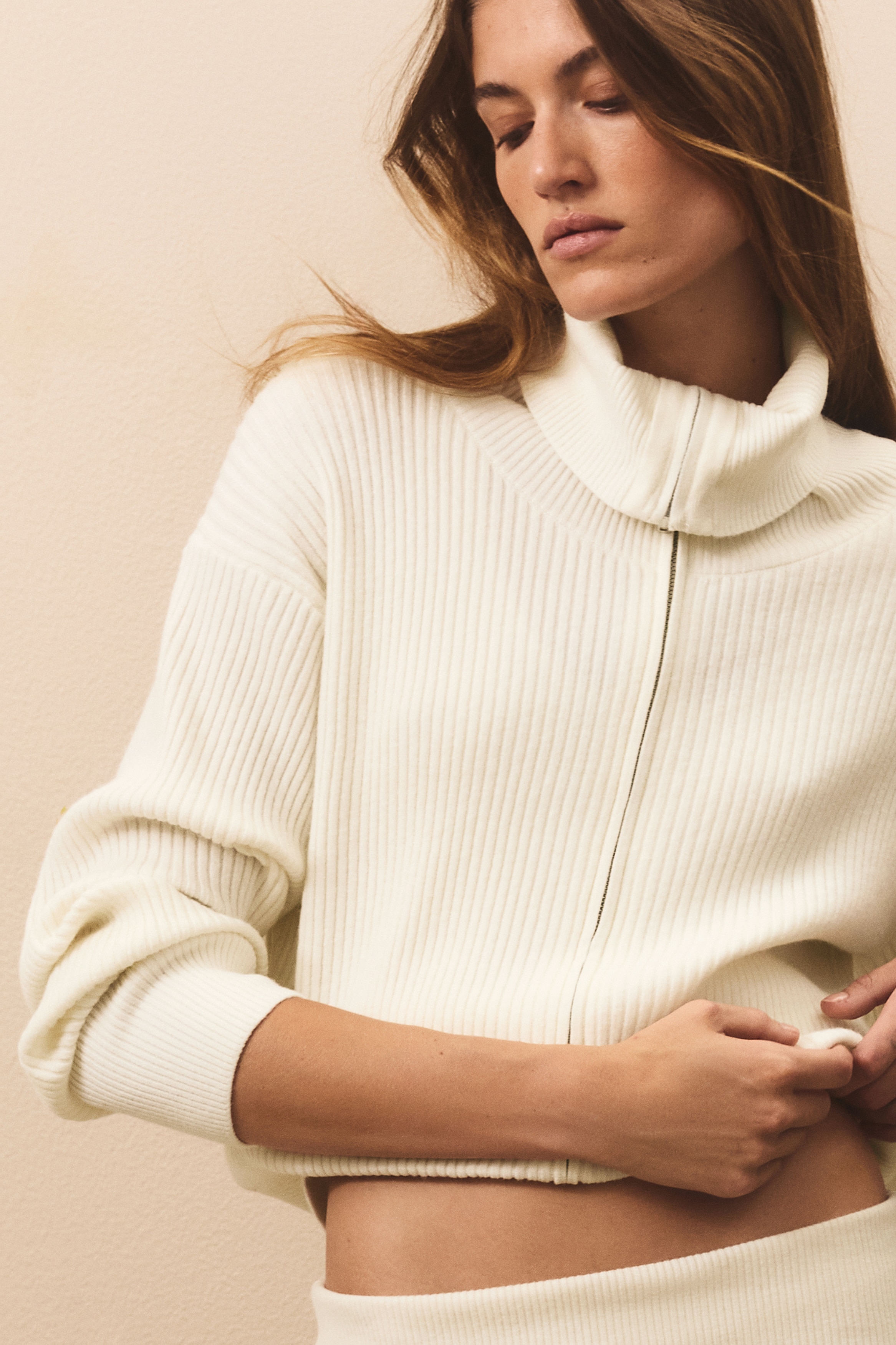 En offwhite ribbestrikket cardigan er vendt mot fronten, med hÃ¸y funnelneck og en heldekkende glidelÃ¥s i front. Cardiganen viser et vertikalt ribbestrikket mÃ¸nster med brede ribbestrikkede mansjetter og en matchende ribbestrikket kant nederst.