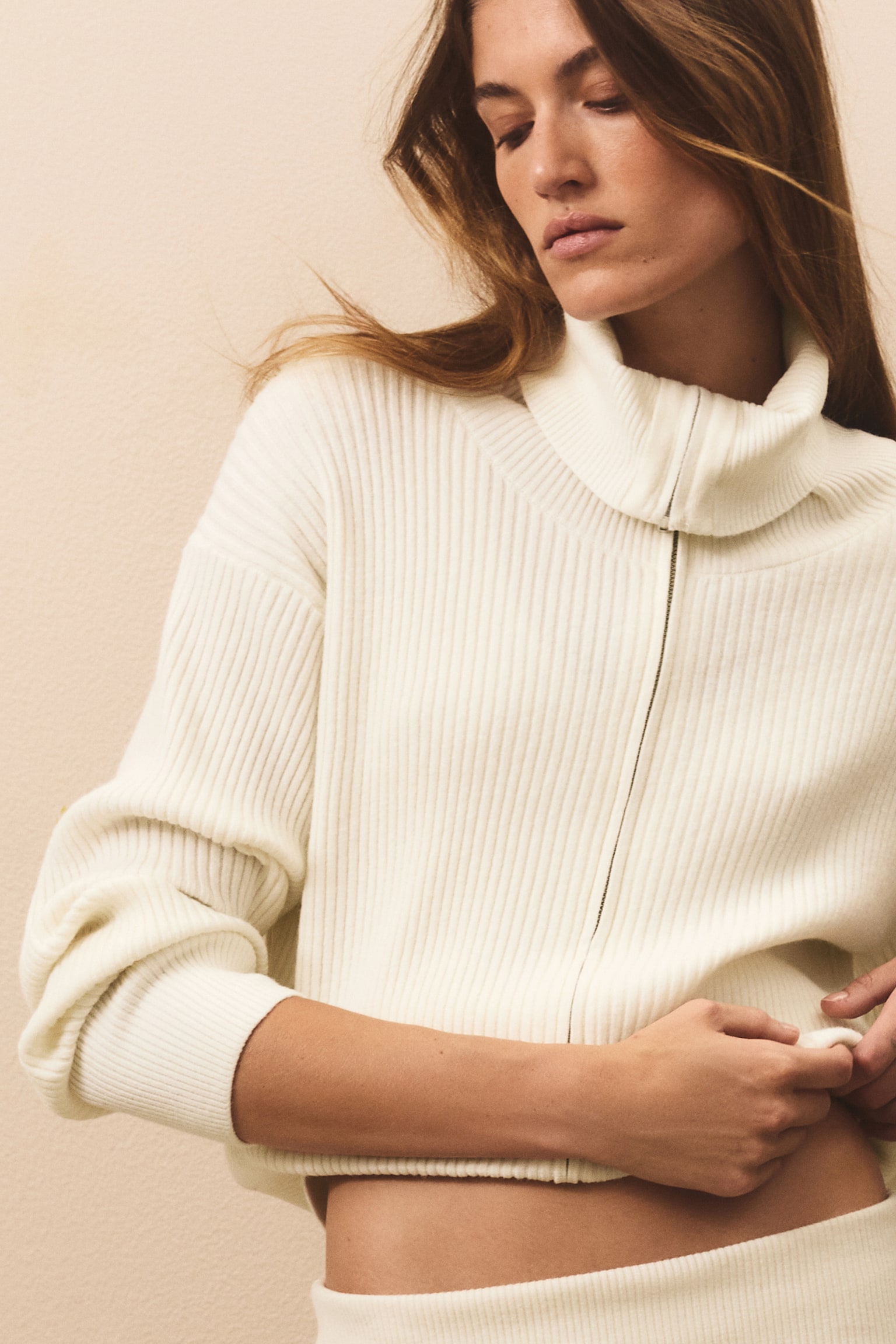 An off-white ribbed cardigan is facing the front, featuring a high funnel neck and a full-length front zip closure. The cardigan is showcasing a vertical ribbed-knit pattern with wide ribbed cuffs and a matching ribbed hem. - 1