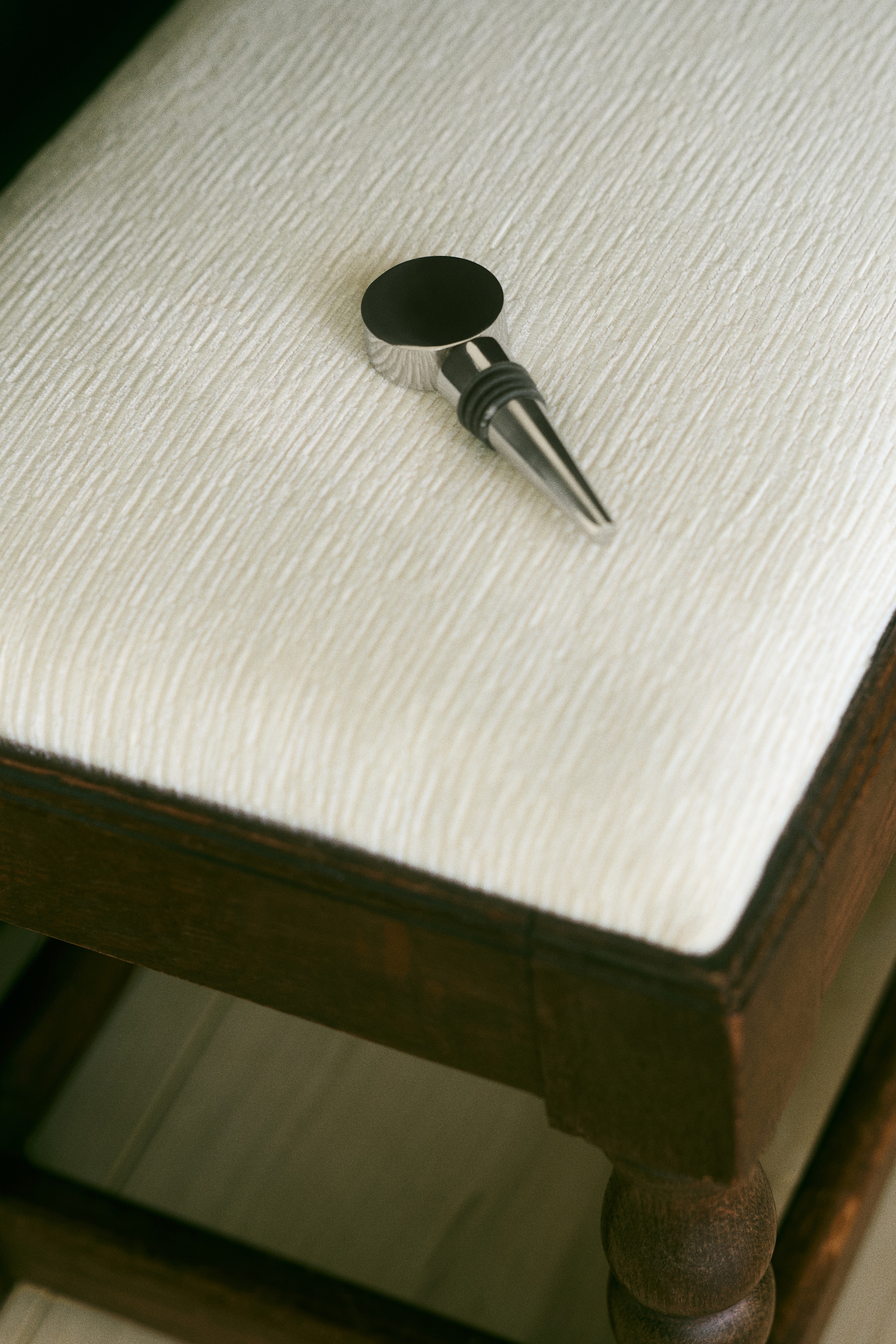 A metallic bottle stopper is featuring a shiny, flat circular top, a cylindrical mid-section, and a tapered conical base with two black rubber rings. It is lying horizontally within an open, light-colored box.