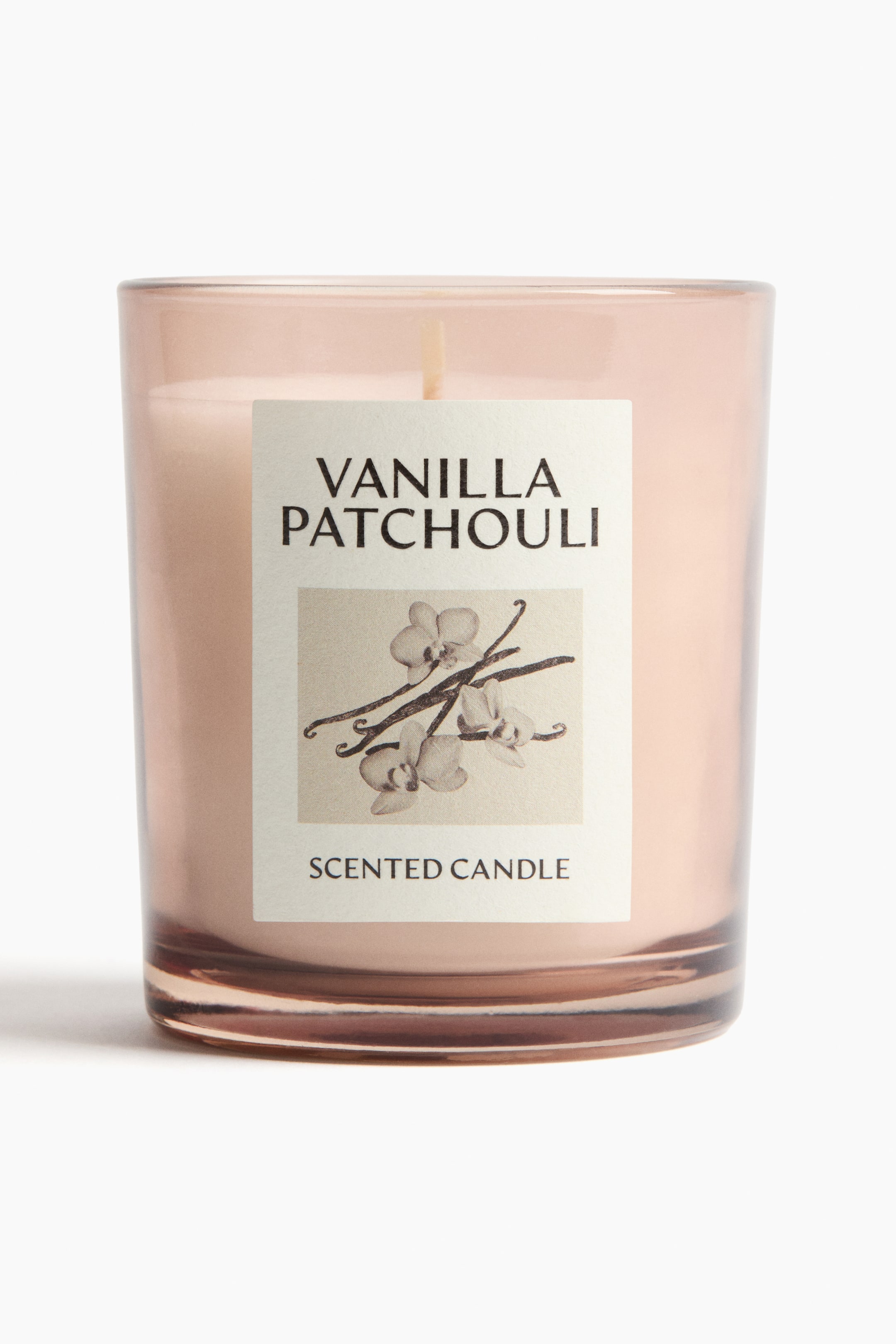 View larger image: A scented candle is presenting its front, encased in a translucent, light pink glass holder. The holder is containing light pink wax with a single, central wick protruding, and a rectangular label is affixed to the front displaying "VANILLA PATCHOULI" and "SCENTED CANDLE" with an illustration of vanilla pods and orchids.
