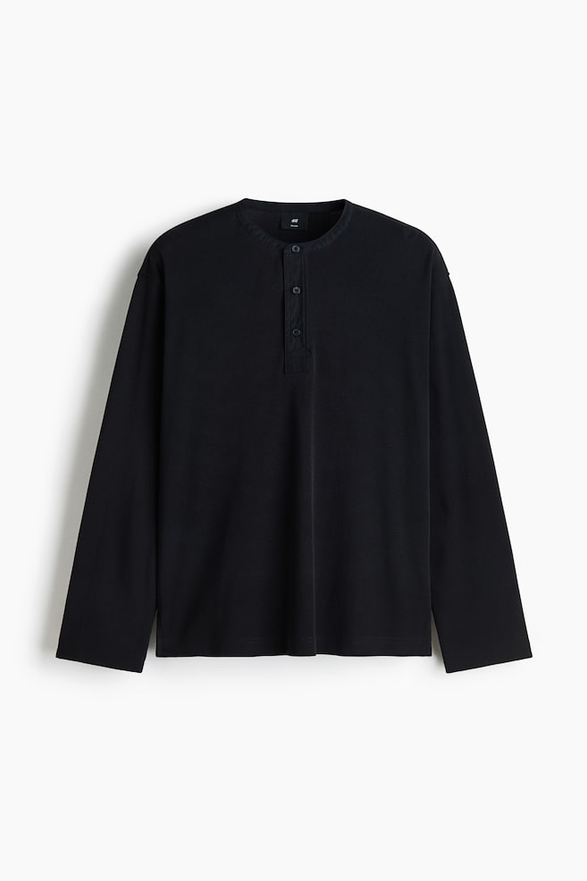 A black long-sleeve henley shirt is shown facing front, featuring a round neckline with a short, three-button placket. The shirt is presenting a relaxed fit with dropped shoulders and straight, open-hemmed cuffs. - 1
