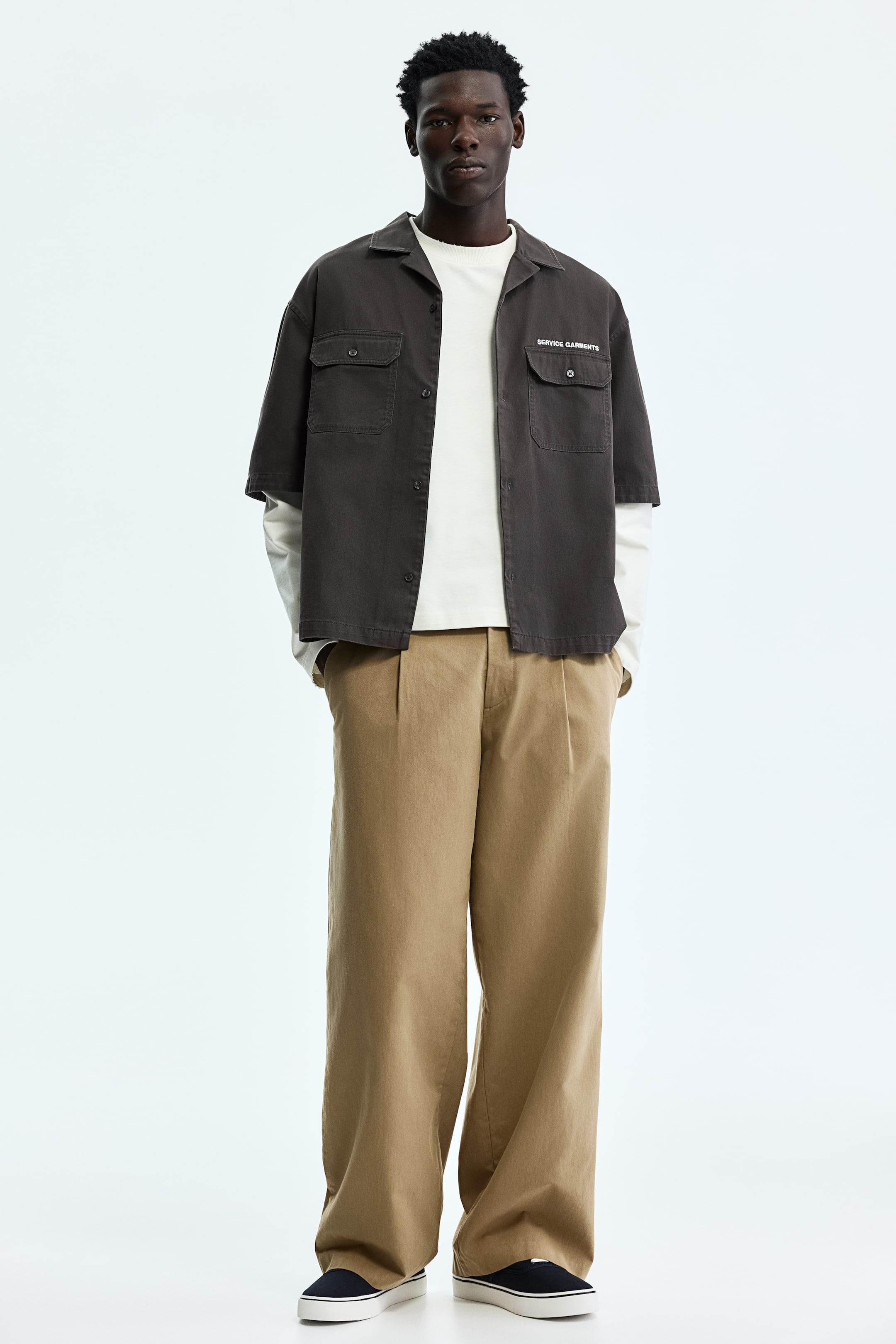 View larger image: A dark brown short-sleeve shirt is facing forward, featuring a camp collar and an open button-up front. It is displaying two chest flap pockets, with "SERVICE GARMENTS" text visible above the left pocket, and is being worn over a white long-sleeve shirt.