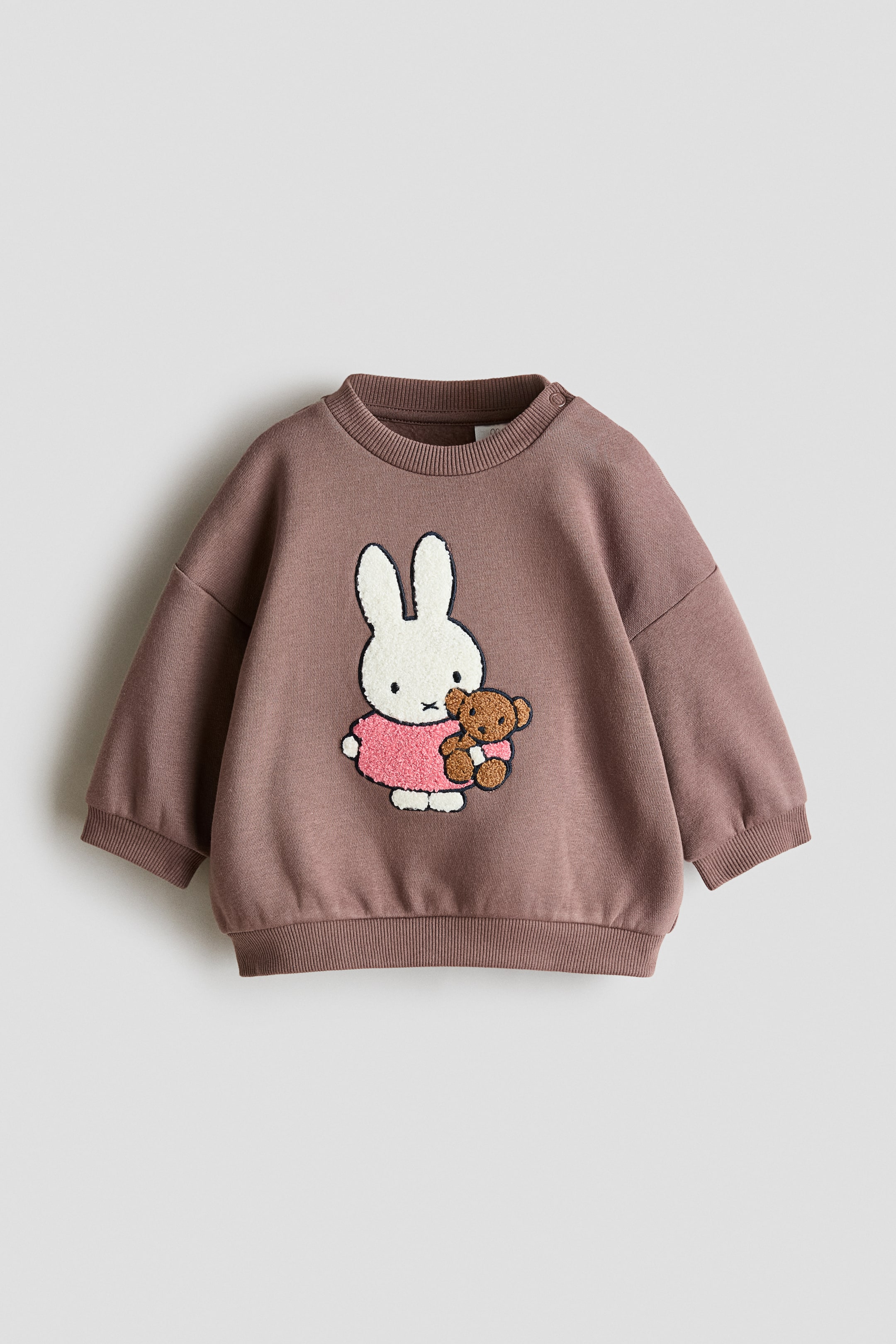 View larger image: A brown sweatshirt is laid flat, facing front. It features a ribbed crew neck with two snap buttons on the right shoulder, ribbed cuffs, and a ribbed hem. A large appliqué of a white bunny wearing a pink textured dress and holding a brown teddy bear is visible on the front.