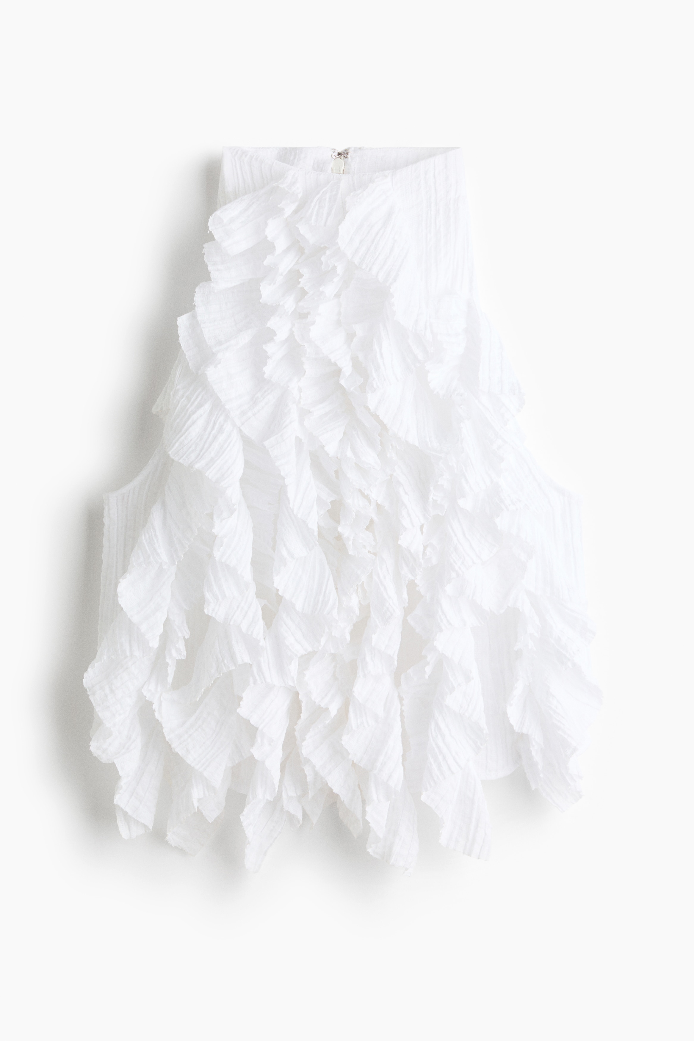 A white sleeveless top featuring cascading pleated ruffles with raw edges and a high round neckline is being worn by the model. A cropped black jacket with braided trim is draped over the model's right shoulder, leaving the other side exposed.
