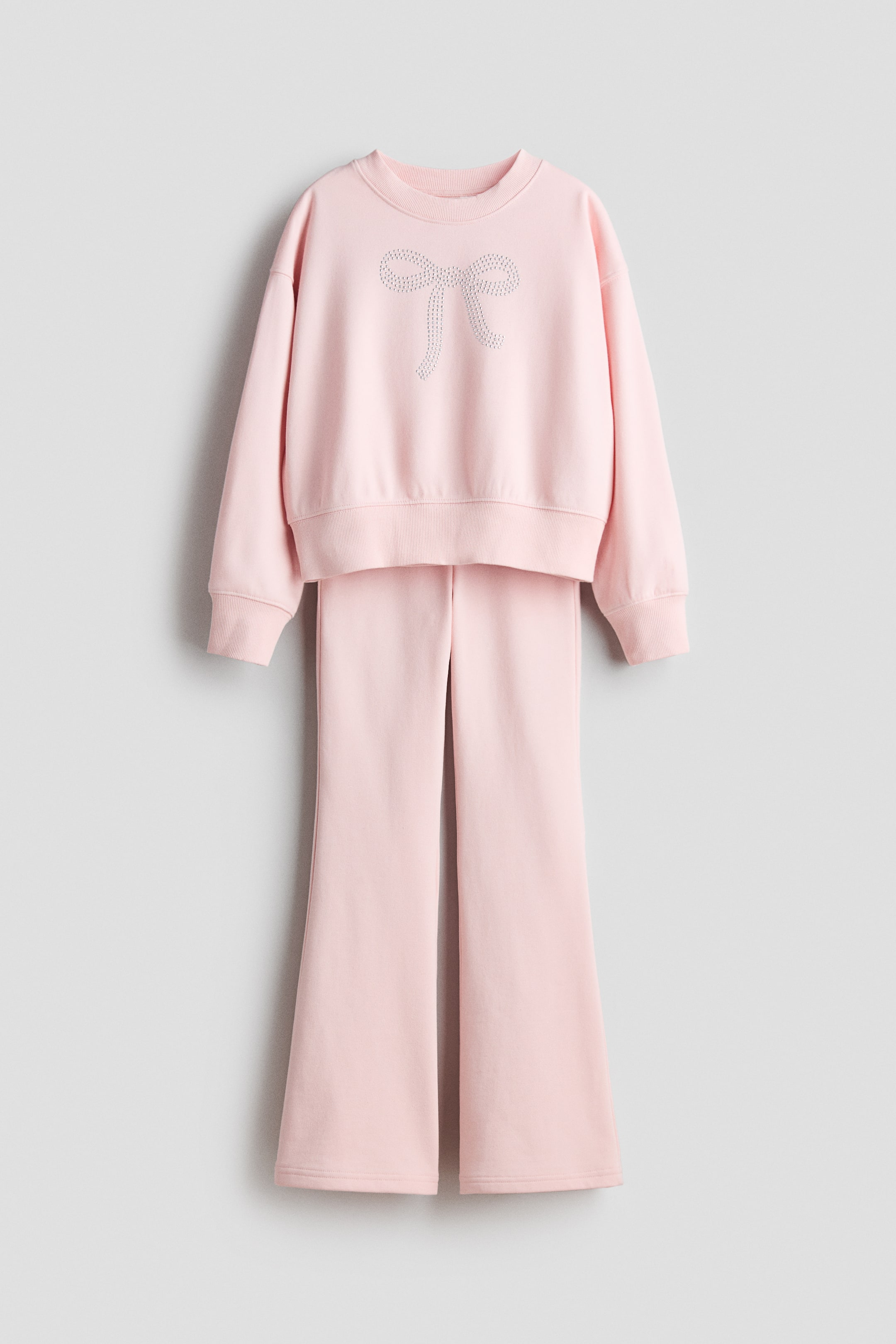 Agrandir l'image: Un ensemble de sweatshirt et jogger rose présente son DEVANT, avec le sweatshirt présentant des épaules tombantes, des détails côtelés au niveau de l'encolure, des poignets et de l'ourlet, et un motif nœud scintillant sur le devant. Les joggers assortis présentent des ourlets larges et évasés.