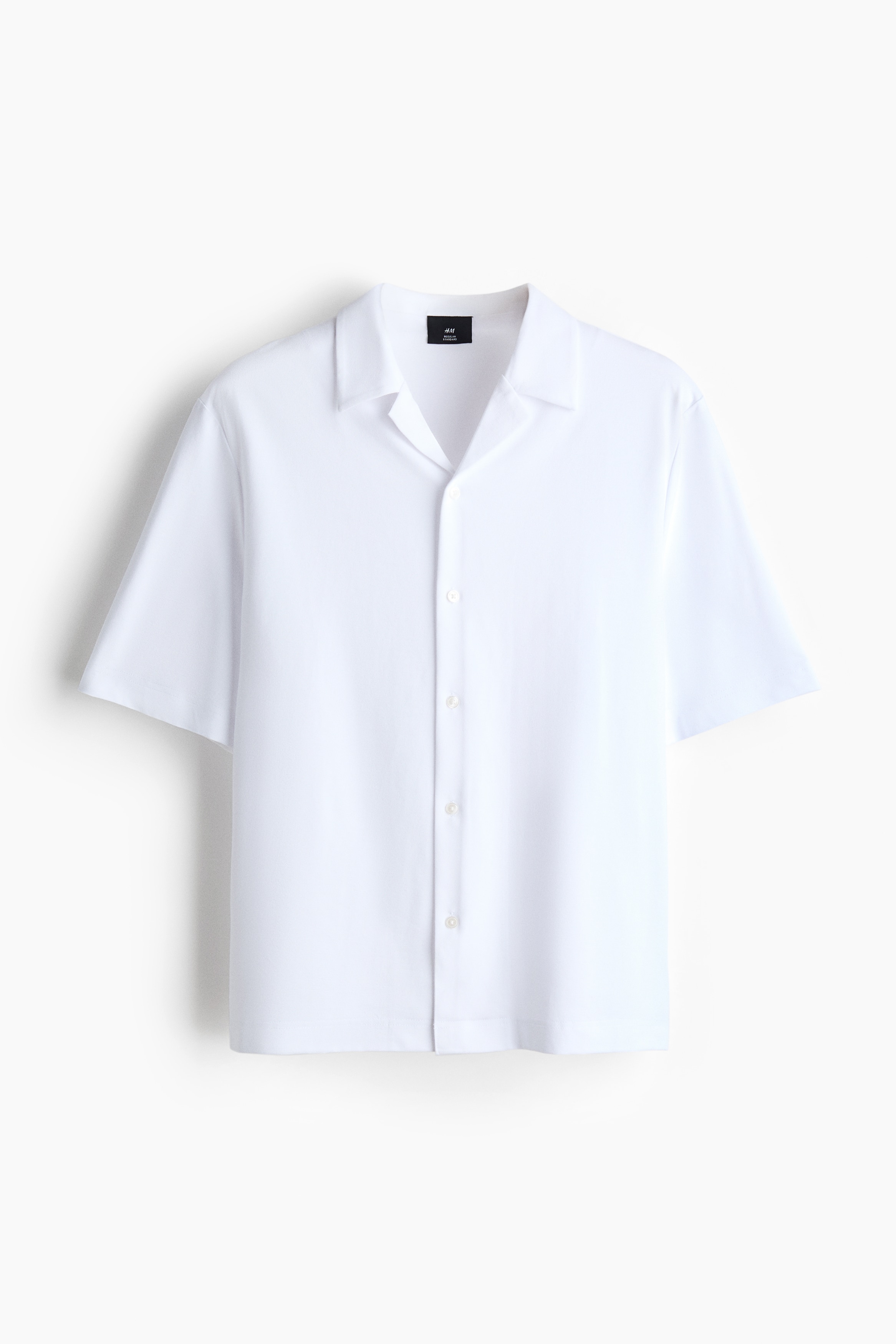 A white short-sleeve shirt is being worn by the model, styled untucked over beige trousers. The shirt features a resort collar, a button-up front, and a straight hem.