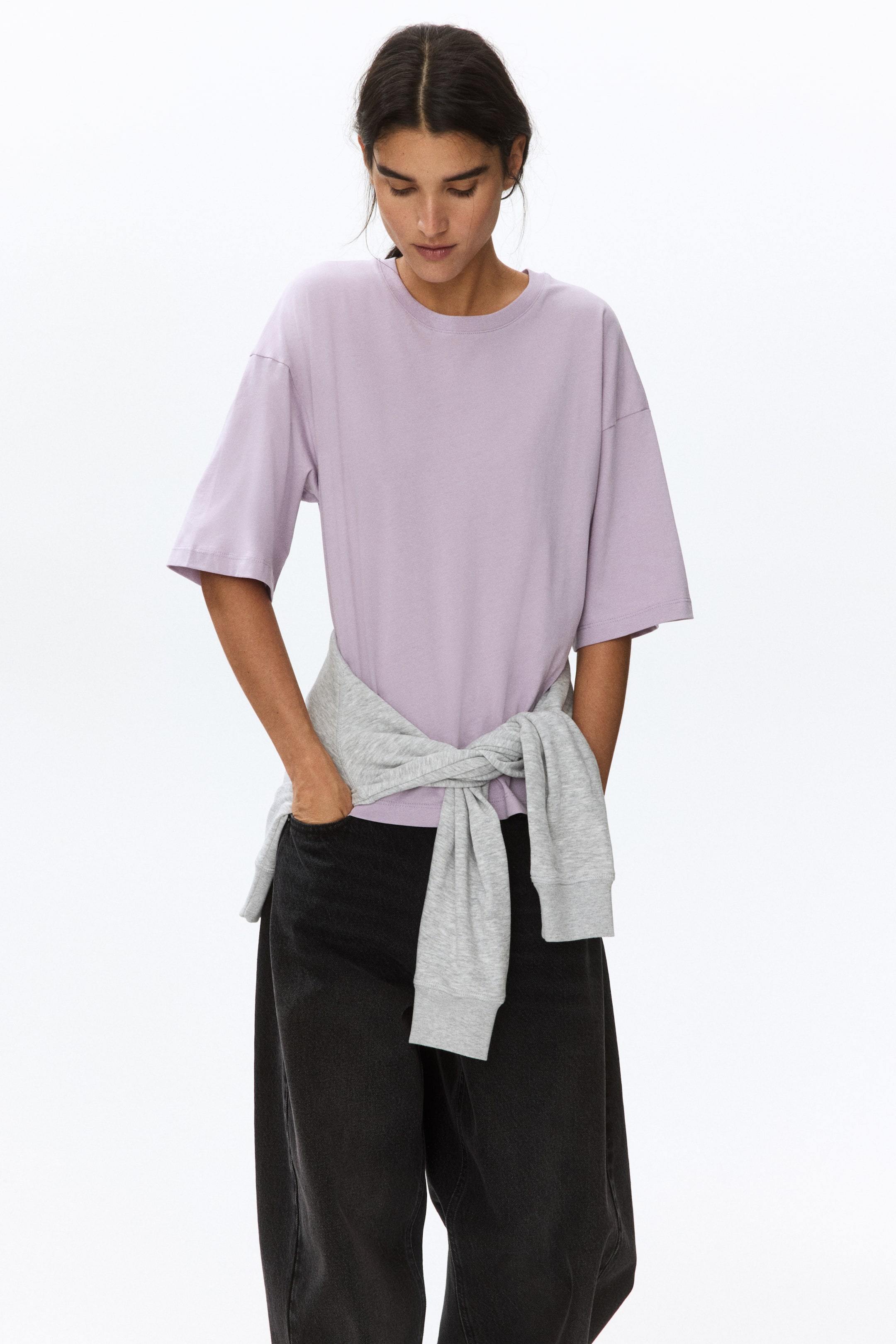 Visa större bild: En ljuslila t-shirt visar upp en avslappnad passform, med en rund hals och korta, vida ärmar. En grå sweatshirt är knuten runt midjan, vilket påverkar dess synliga längd.