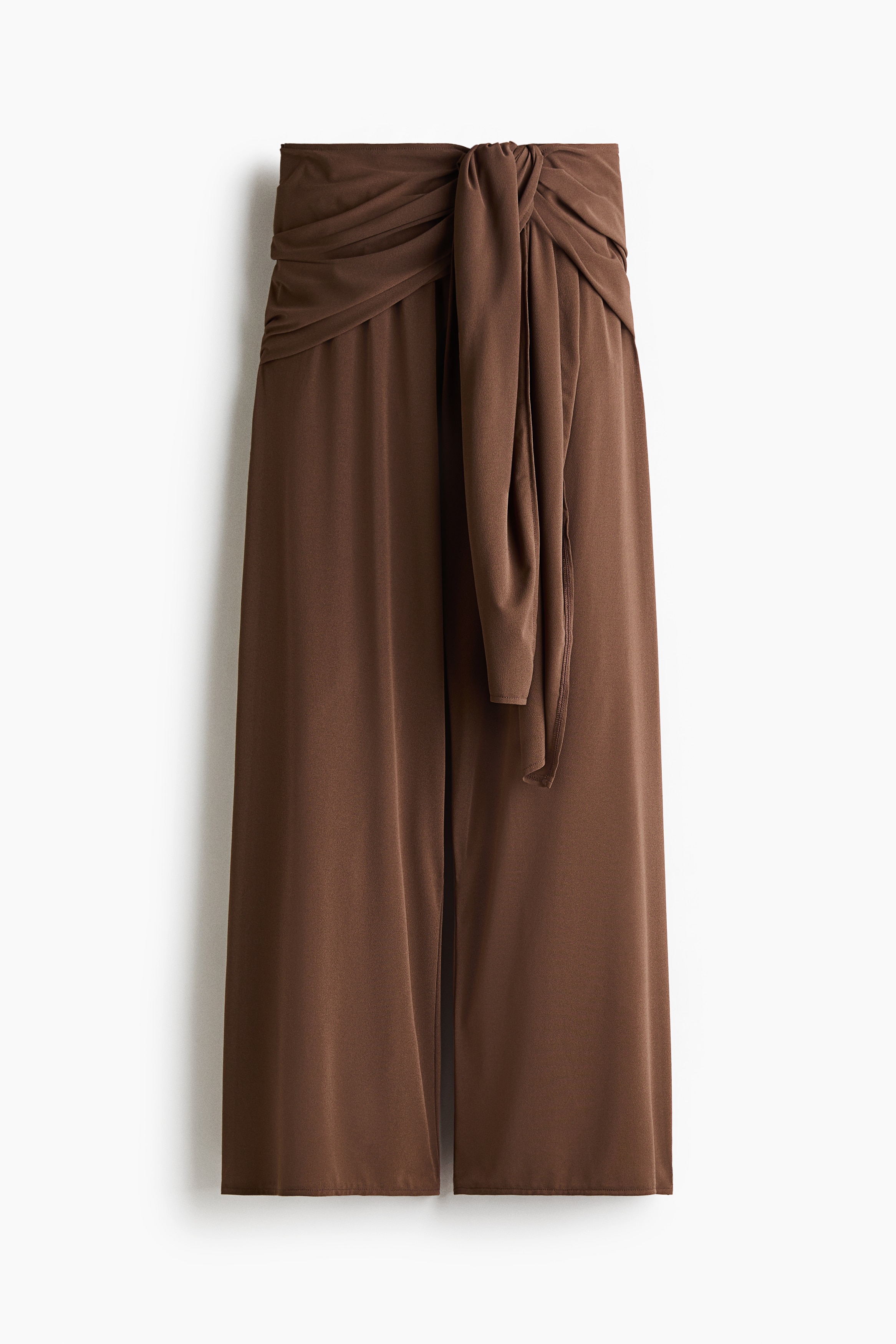 The model is wearing brown textured jersey trousers, featuring a relaxed fit, a high waist, and wide side panels that drape and tie at the front. The wide-leg trousers are paired with a matching brown cropped top with a high neck and cold-shoulder cut-outs.