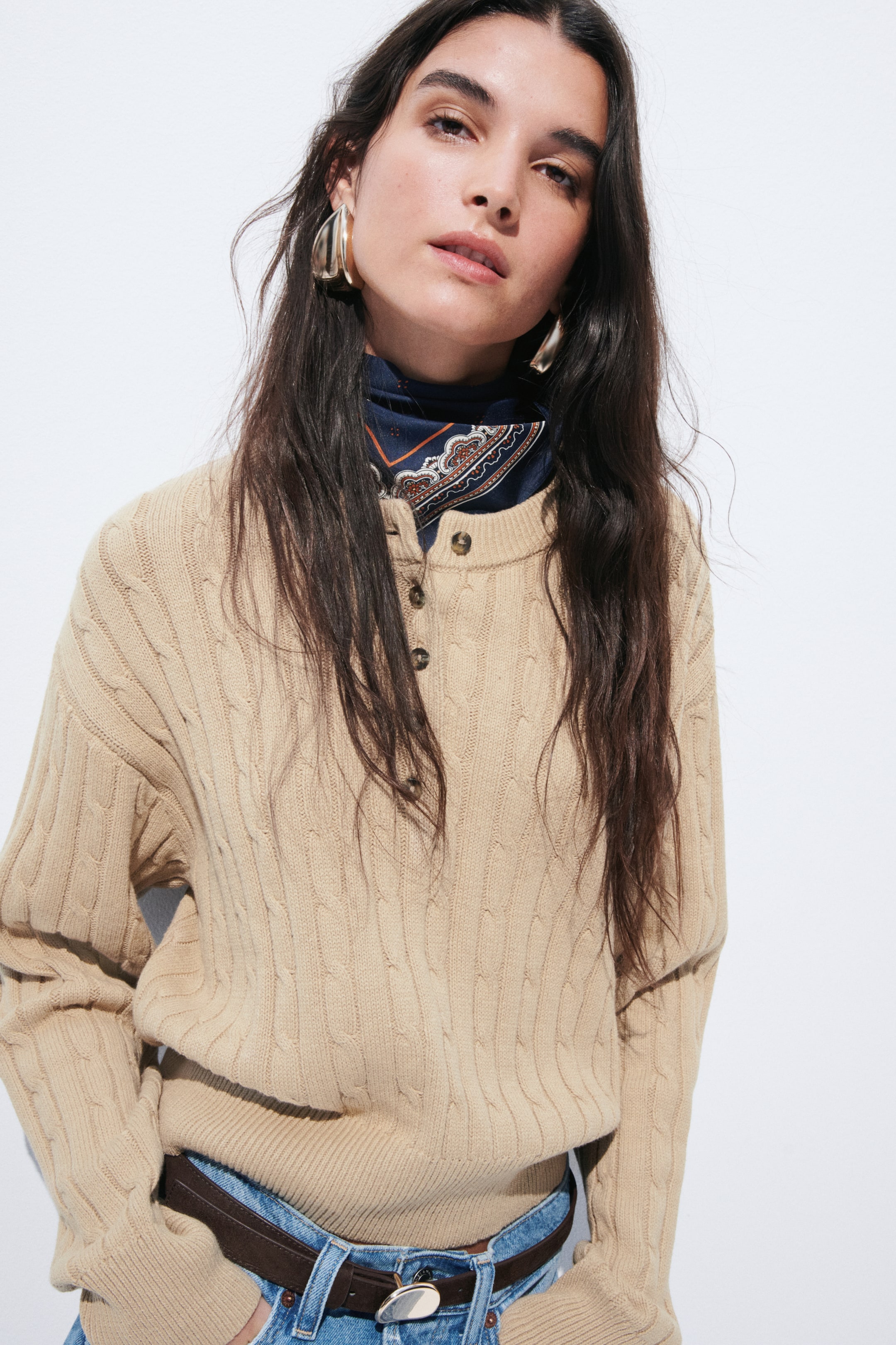 View larger image: A beige cable-knit jumper features a crew neck with a three-button henley placket, showcasing a distinct cable-knit pattern, ribbed cuffs, and a ribbed hem. A dark blue patterned scarf is visible beneath its neckline, and its hem is tucked into blue jeans.