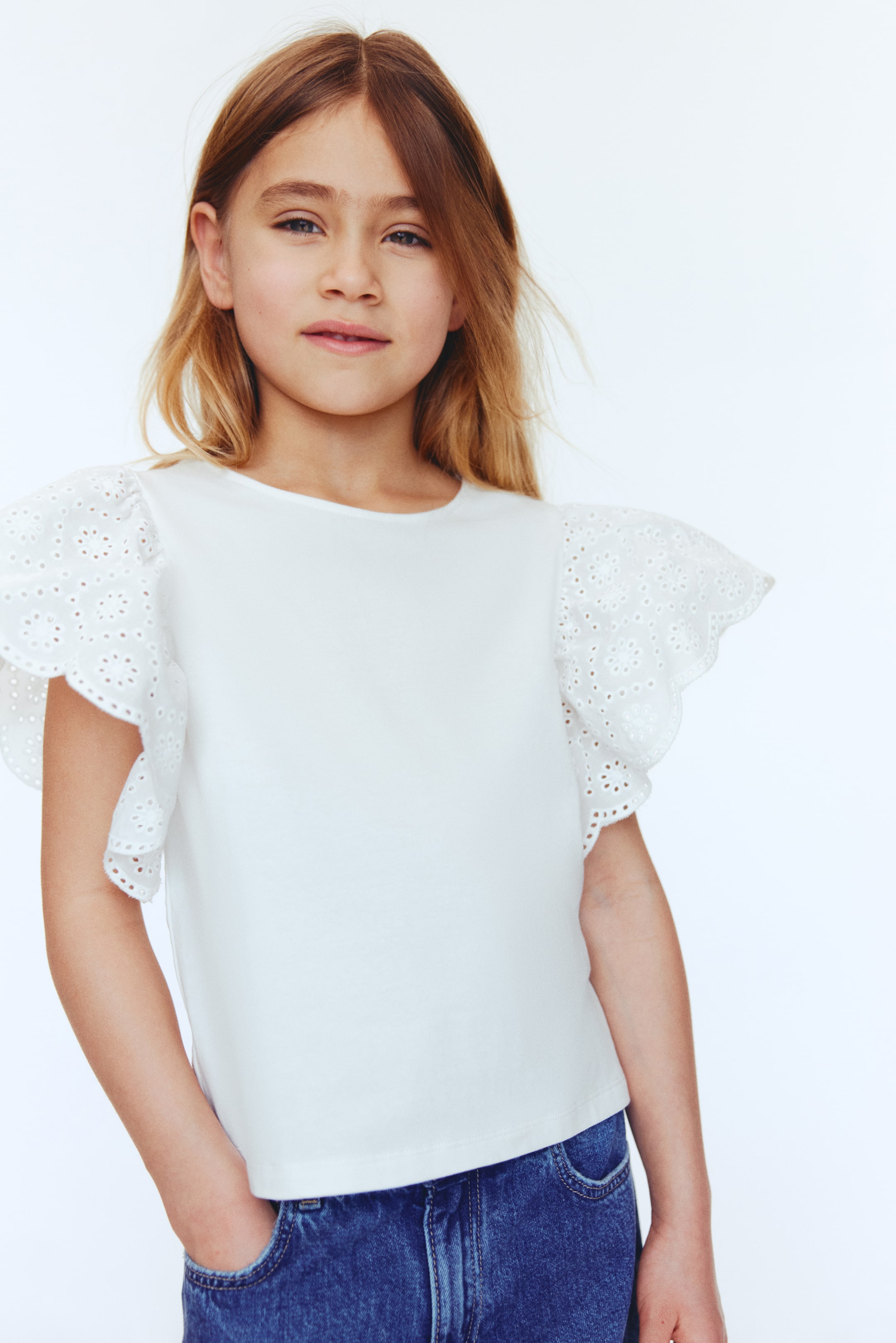 Ampliar la imagen: Una blusa blanca con mangas de vuelo bordadas con ojales y bordes festoneados, y cuello redondo. Se lleva sobre jeans de mezclilla azul oscuro.