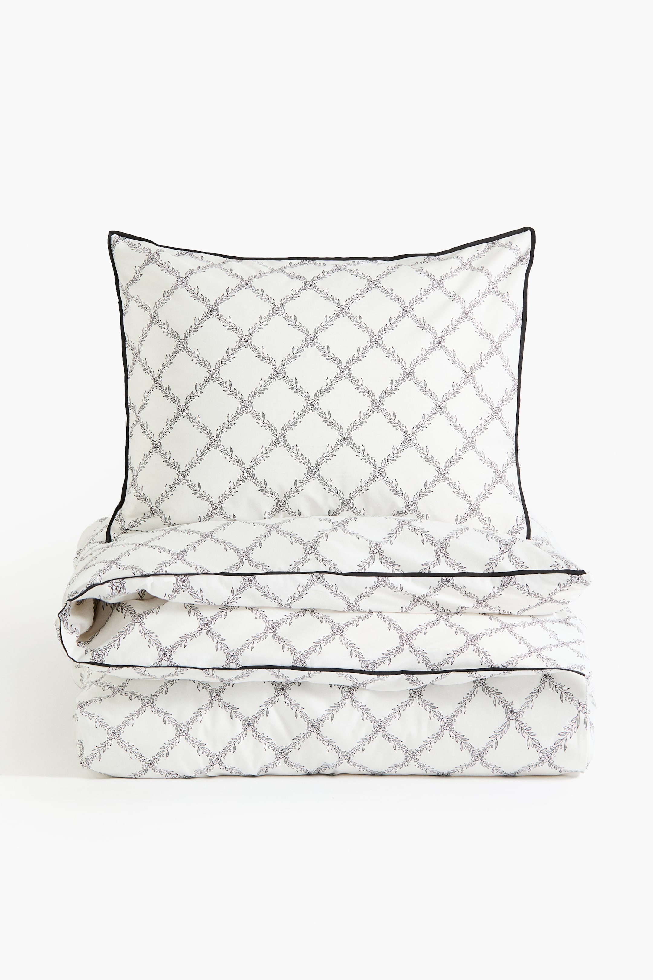 Größeres Bild ansehen: Das Bettwäsche-Set wird frontal präsentiert: ein gefalteter Bettbezug mit einem darauf gestapelten Kissenbezug. Beide Teile weisen eine weiße Grundfarbe mit einem flächendeckenden dunklen Gittermuster aus blättrigen Ranken sowie eine schwarze Paspelierung an den Kanten auf.