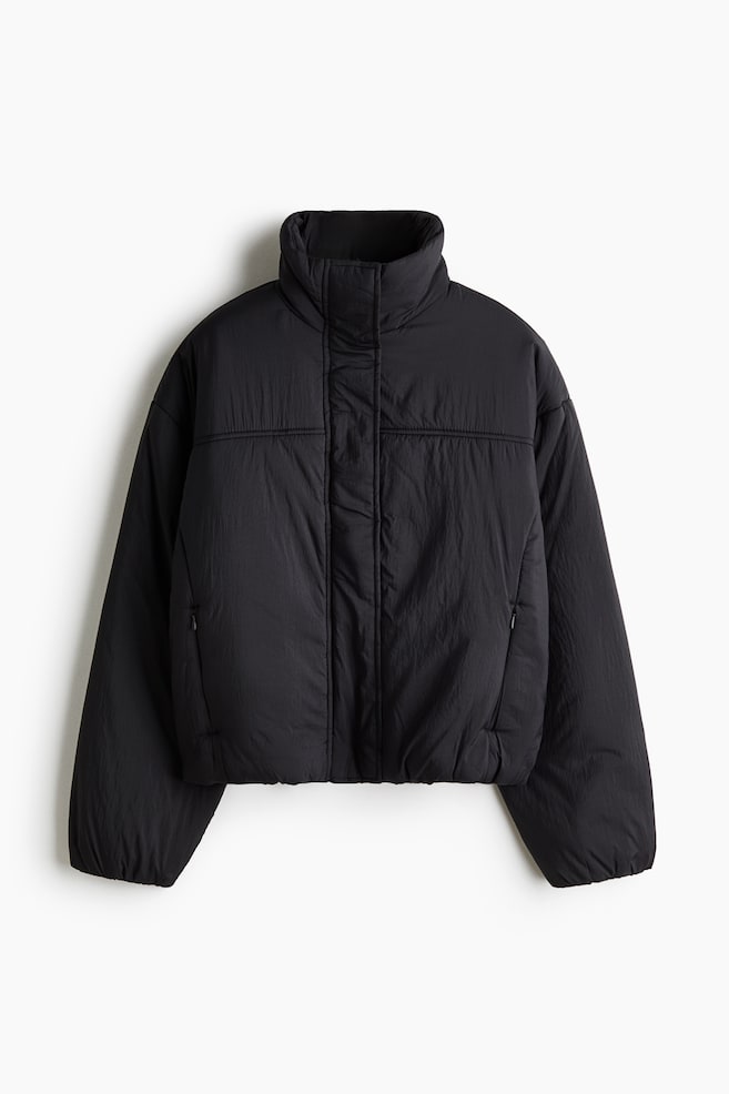 An oversized, padded black nylon jacket is shown from the front, featuring a high stand collar, a concealed front fastening with a wind flap, and a horizontal chest yoke. The jacket is designed with dropped shoulders, long sleeves with elasticated cuffs, and discreet zippered side pockets. - 1