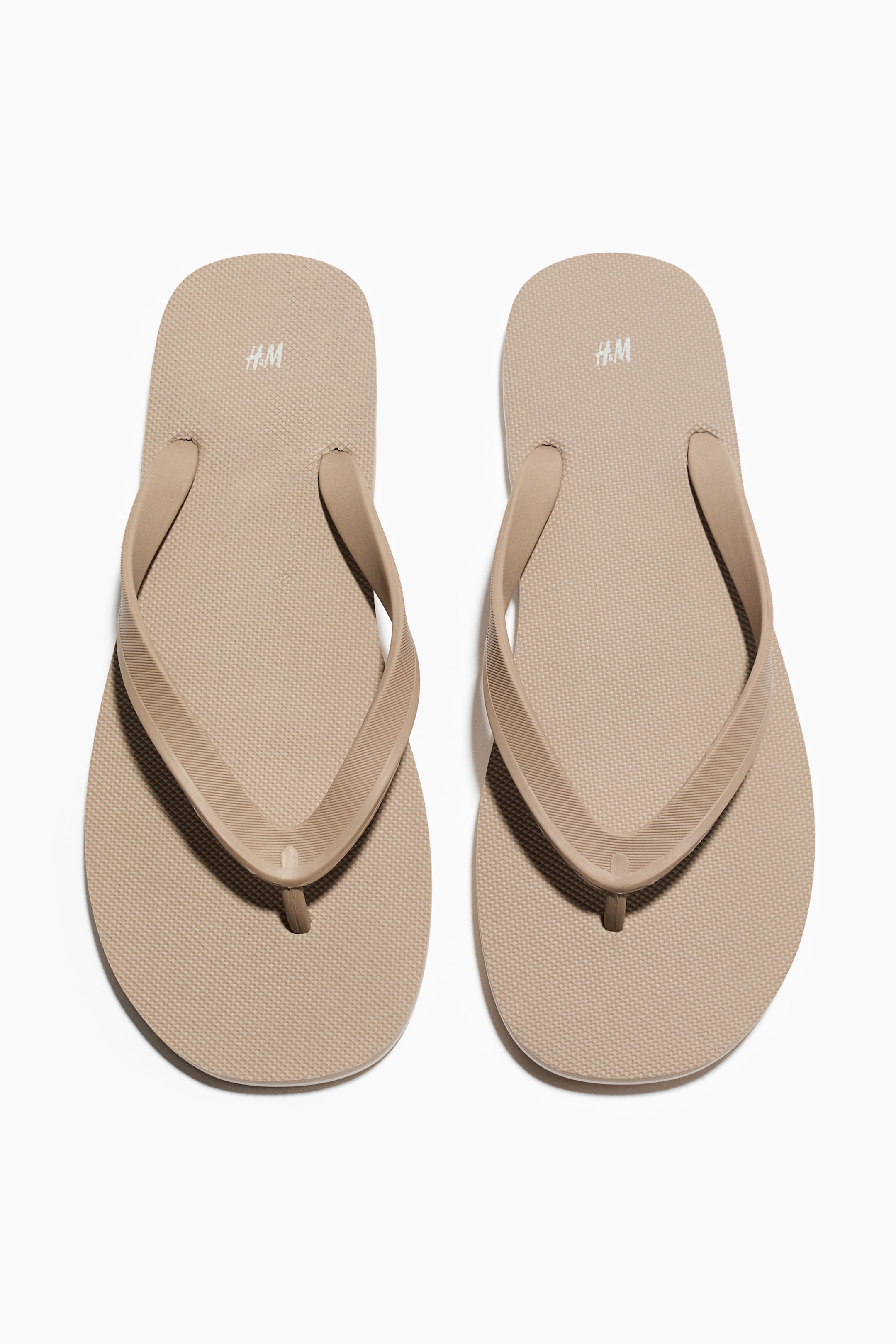 Visa större bild: Ett par beige gummiflipflops med remmar och en smal tårem presenteras från en framåtvänd, uppifrån och ner vy. Varje flipflop visar en texturerad sula och texturerade remmar, med en liten "H.M"-logotyp synlig nära hälen.