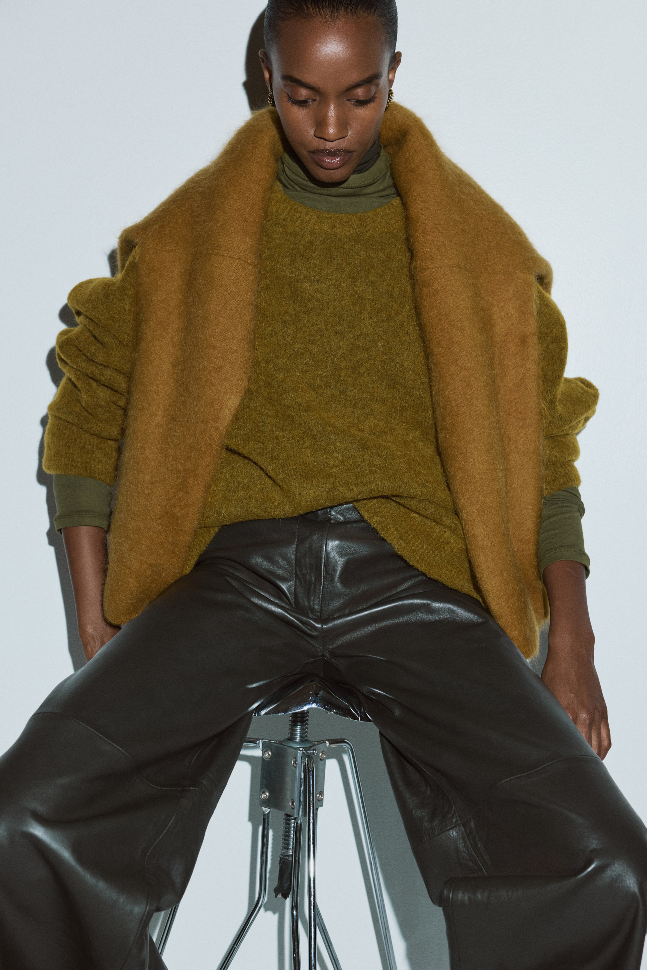 Agrandir l'image: Un pull en maille épaisse vert olive, avec une surface texturée et des manches volumineuses, est porté sur un col roulé vert olive. Un châle duveteux couleur camel est drapé sur les épaules, et un pantalon en cuir marron foncé à jambes larges avec des coutures visibles est porté.