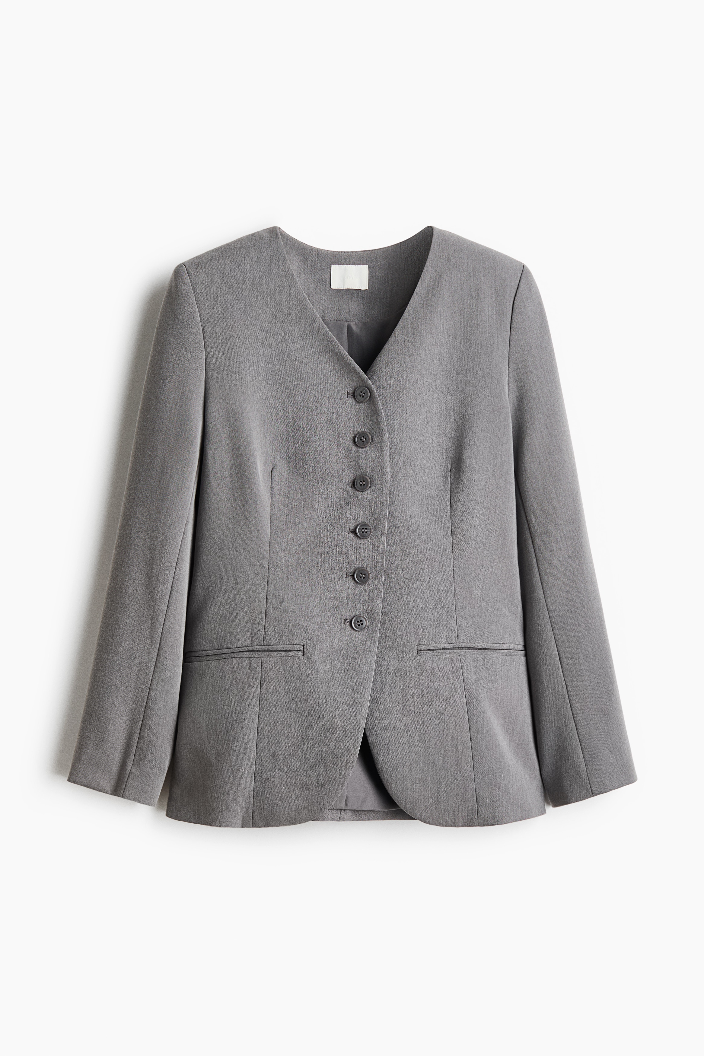A grey lapelless twill blazer featuring a deep V-neckline, front buttons, and jetted pockets is worn by the model, who stands with her arms crossed. The blazer is styled with high-waisted black wide-leg jeans and black pointed-toe heeled boots.