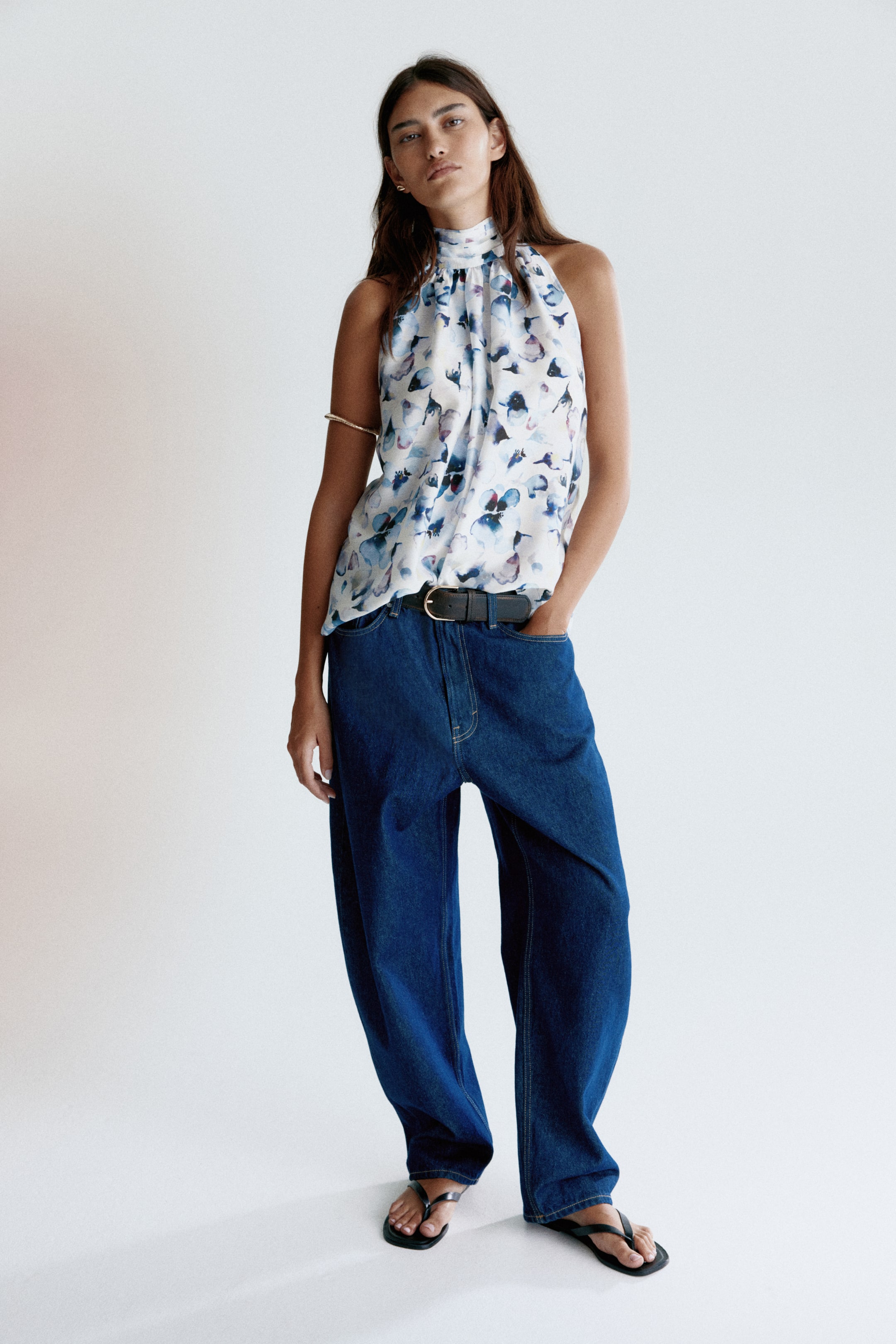 View larger image: A white halter neck top is facing front, displaying an abstract blue, purple, and black floral print. It is featuring a gathered high neckline and is tucked into dark blue jeans.