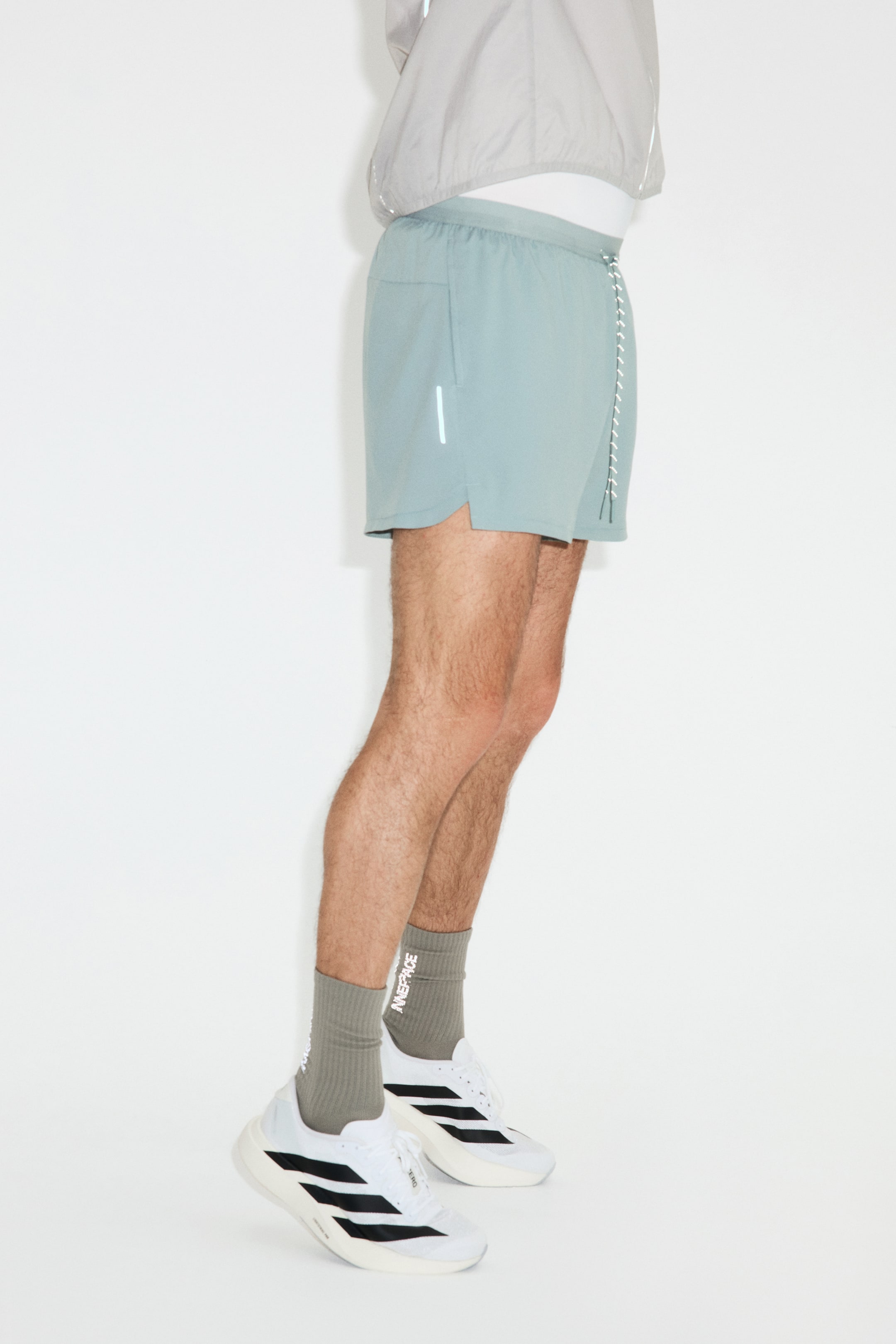 View larger image: Seafoam green running shorts are showcased from a side-front angle, featuring an elastic waistband with a white drawstring on the right side, a subtle side slit pocket on the left, and a short reflective vertical stripe on the lower left leg.