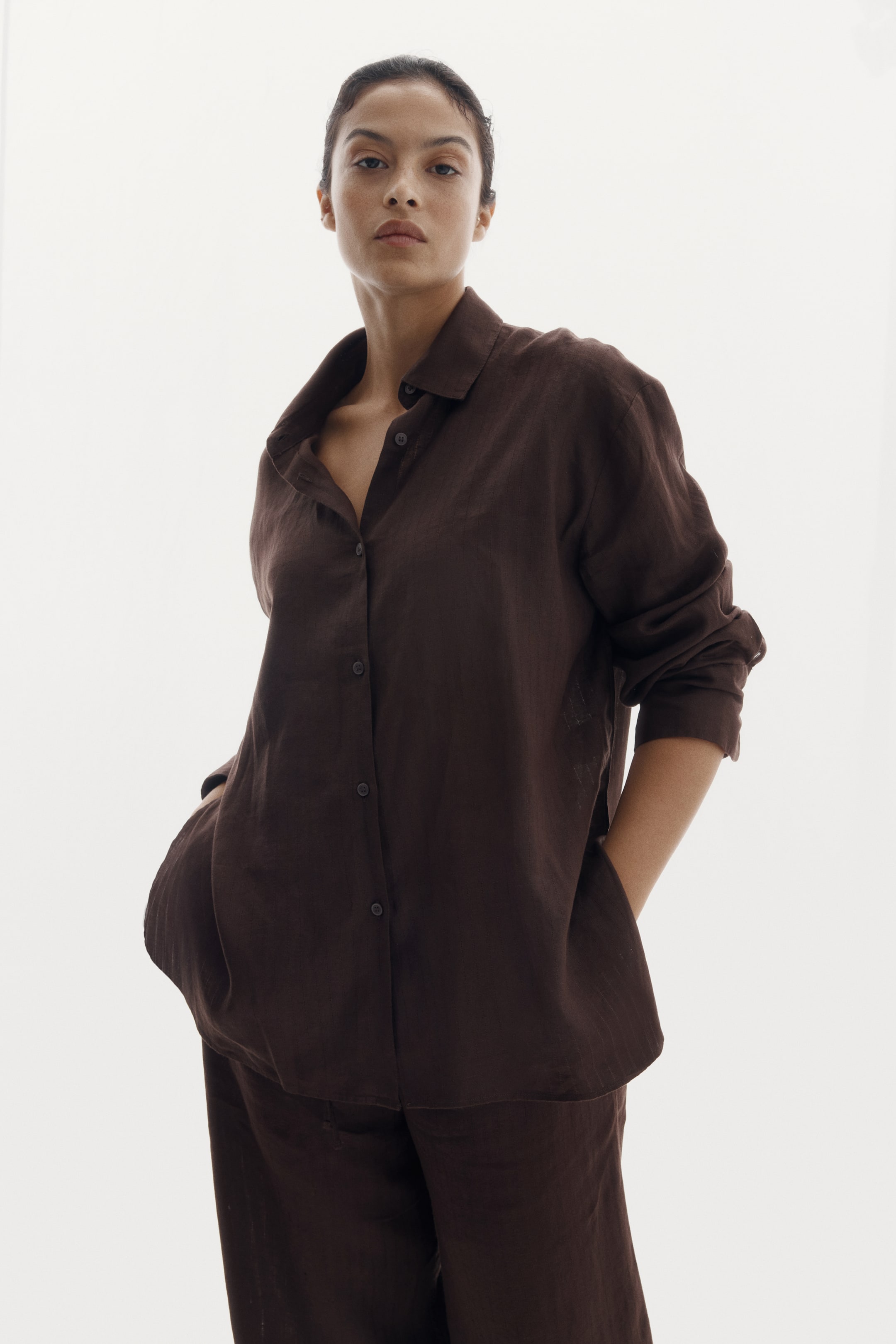 View larger image: "A dark brown long-sleeved shirt with a classic collar and button-down front is being worn, draping loosely over matching relaxed trousers, with hands in the trouser pockets."