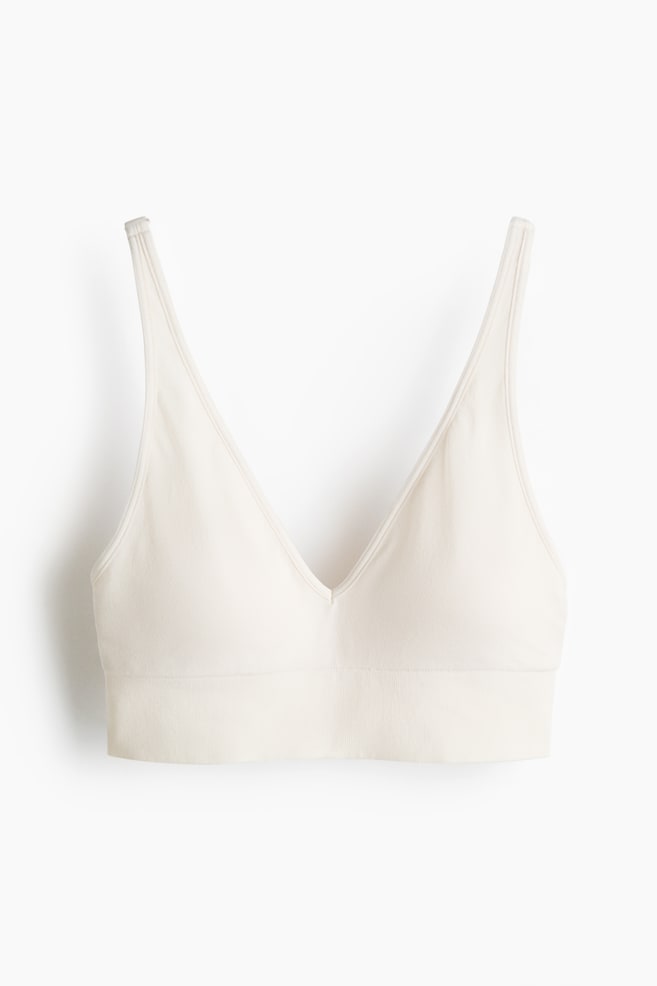 A light beige soft bra is facing front, presenting a v-neckline and thin shoulder straps. It is featuring minimal visible seams, a wide ribbed band at the hem, and no visible fasteners. - 1