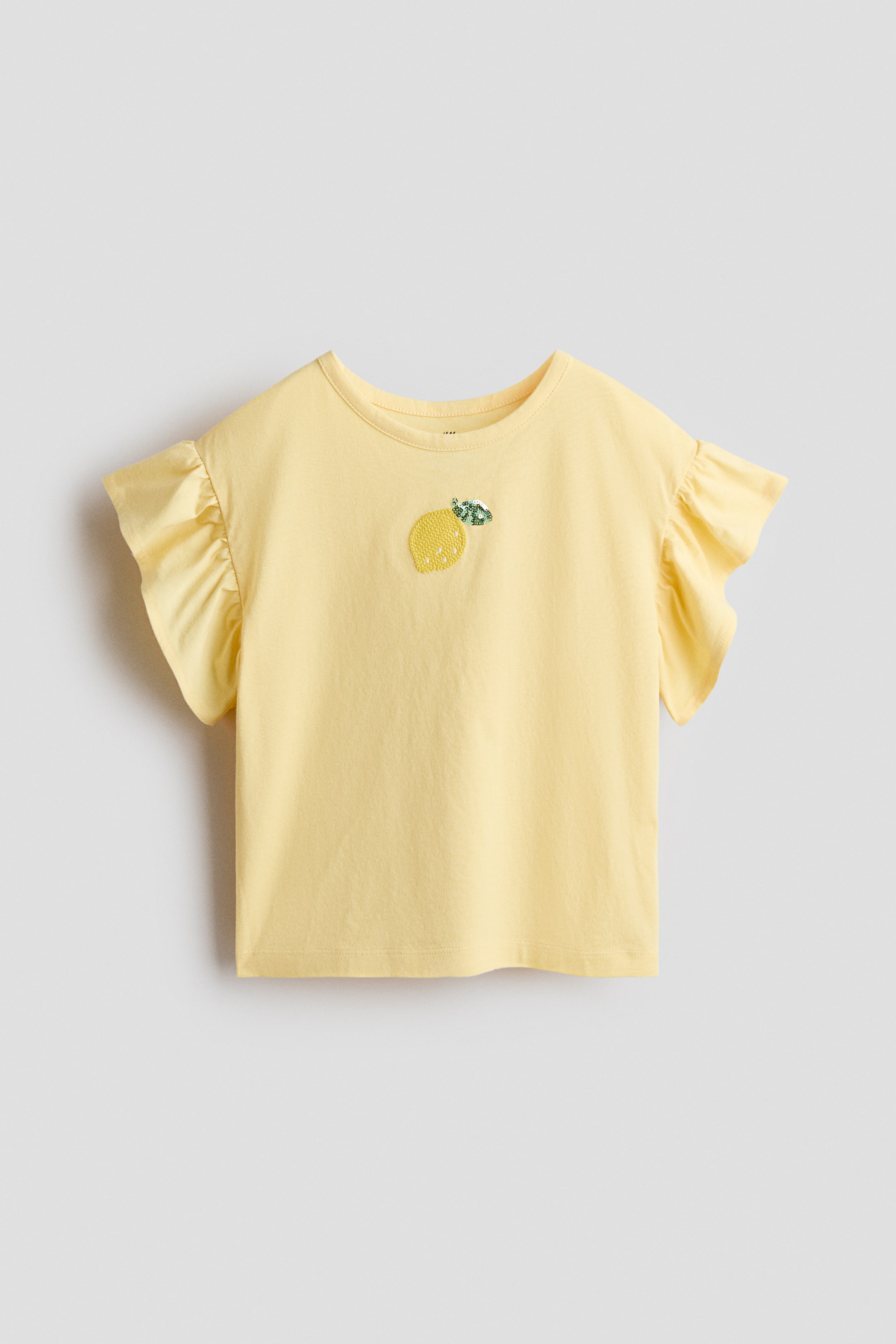 Ver imagem maior: Uma t-shirt amarela clara apresenta uma gola redonda e mangas curtas com folhos. Um motivo de limão com lantejoulas e folhas verdes adorna a frente da camisa.