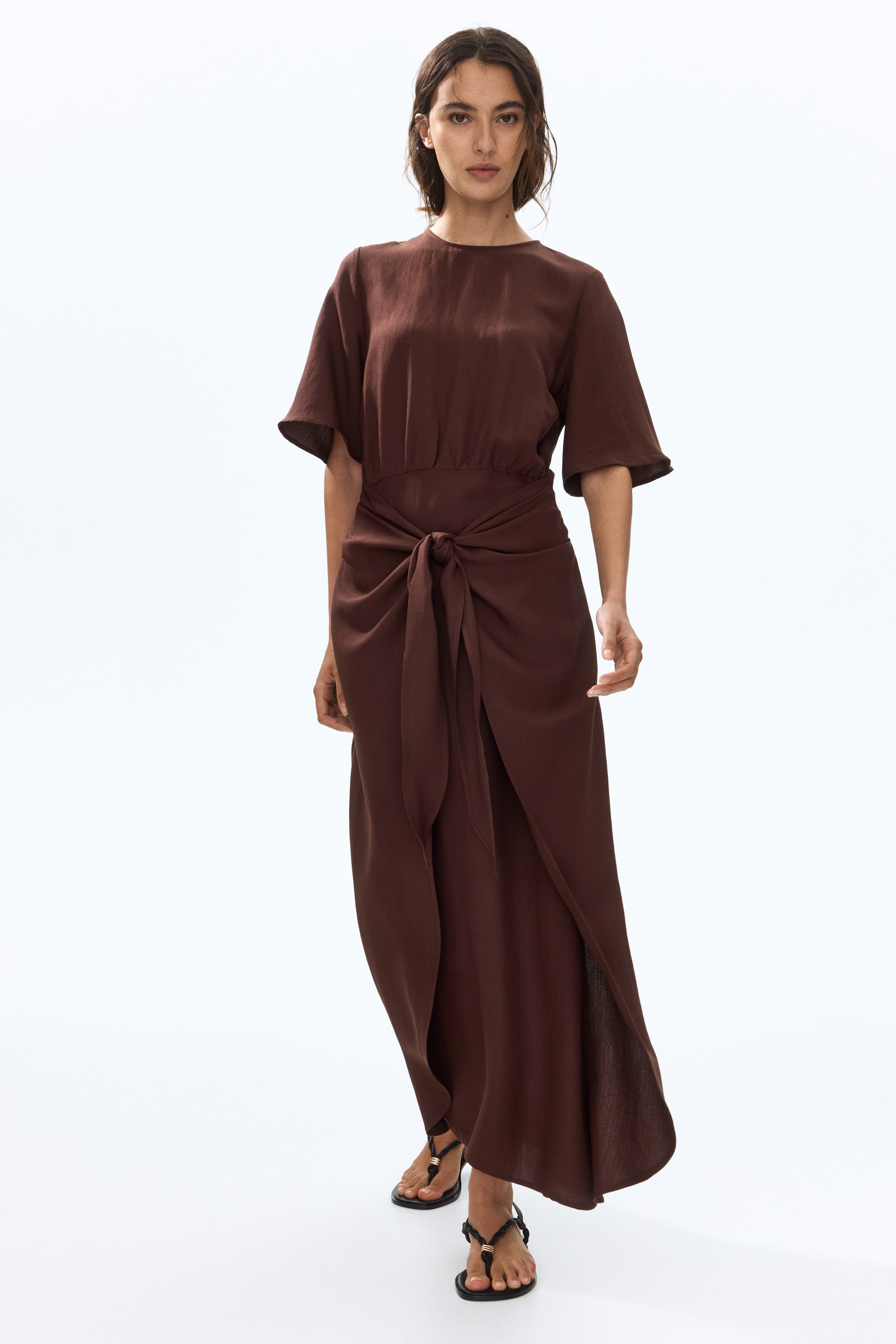 View larger image: The model is showcasing a long brown dress crafted from a viscose-blend weave. It boasts a round neckline, short sleeves, and a gathered waist accentuated by wide ties knotted at the front. A prominent high slit is noticeable at the back of the hem.
