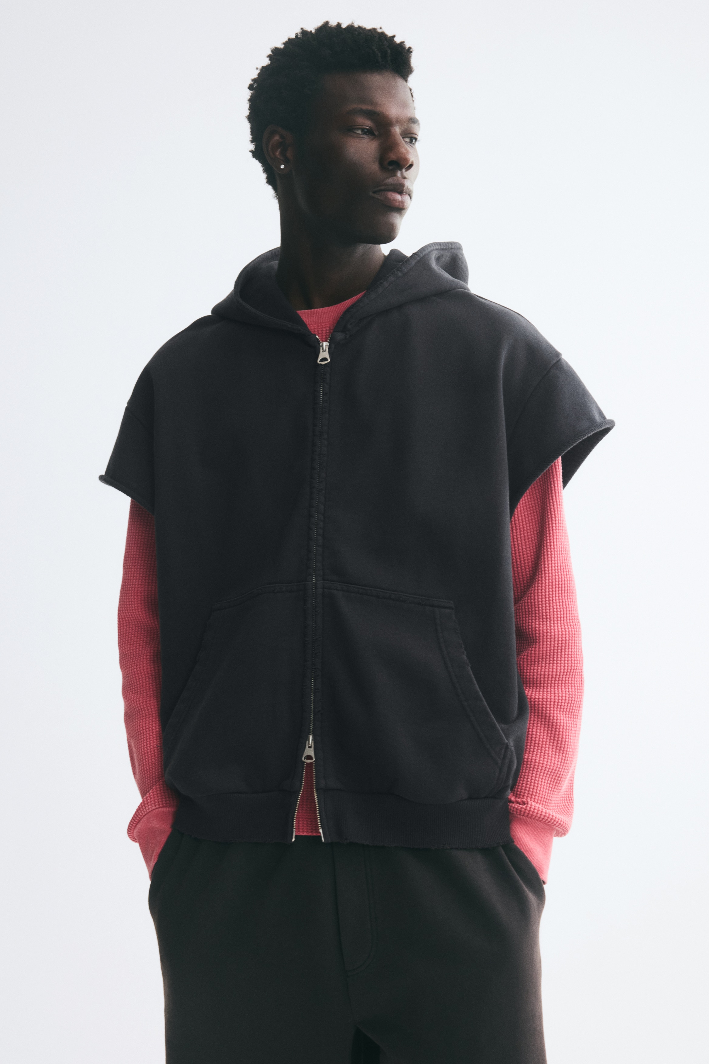 A black sleeveless zip-up hoodie is shown from the front, featuring an attached hood, a full-length front zipper, and a split kangaroo pocket. The garment features rolled-edge armholes and a ribbed hem, which structure its relaxed silhouette.