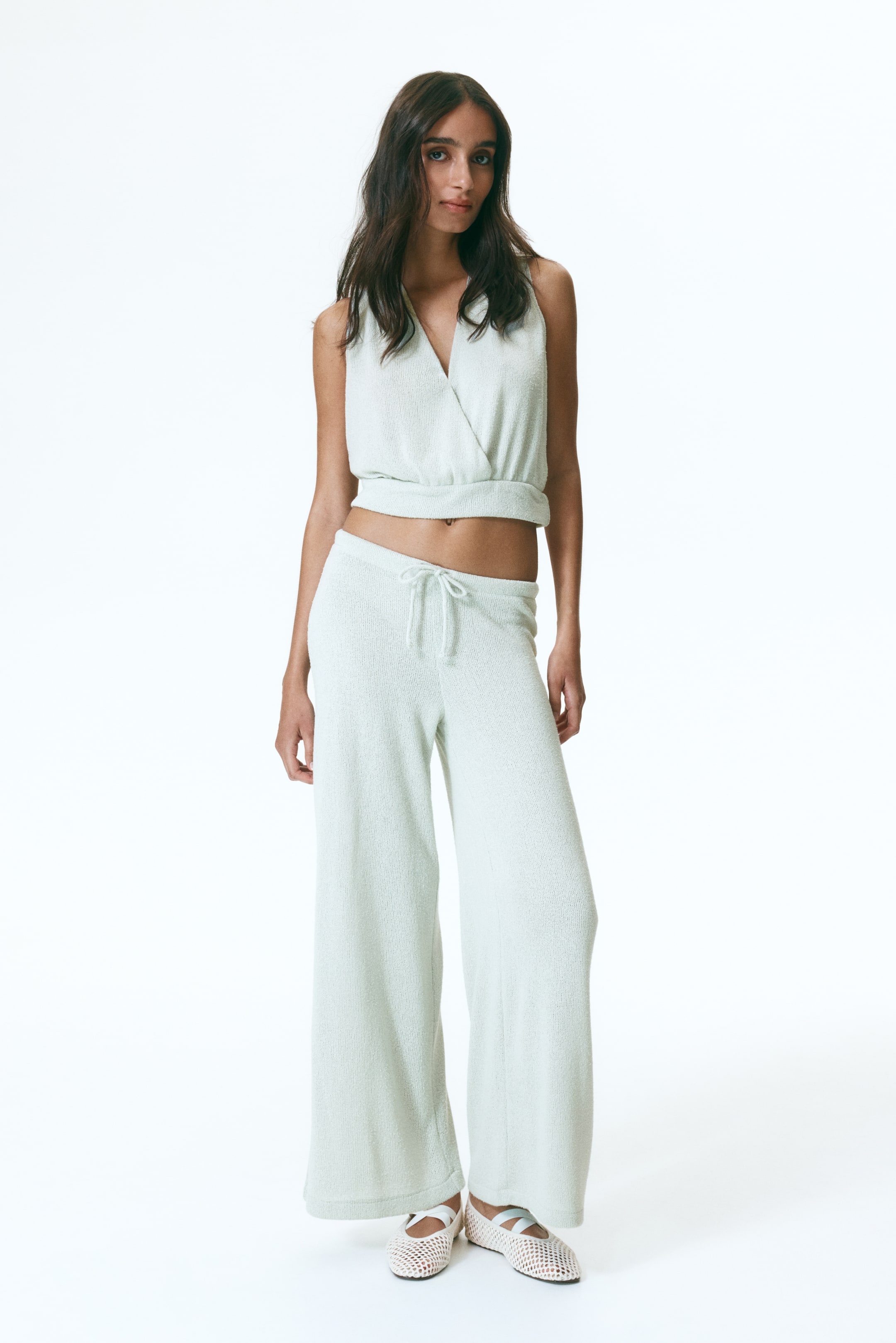 View larger image: Presenting a light green two-piece loungewear set, shown front-facing. This ensemble includes a sleeveless, V-neck wrap-style crop top with an elasticized hem, paired with matching wide-leg pants featuring a drawstring waist. Both pieces are crafted from a soft, textured knit.