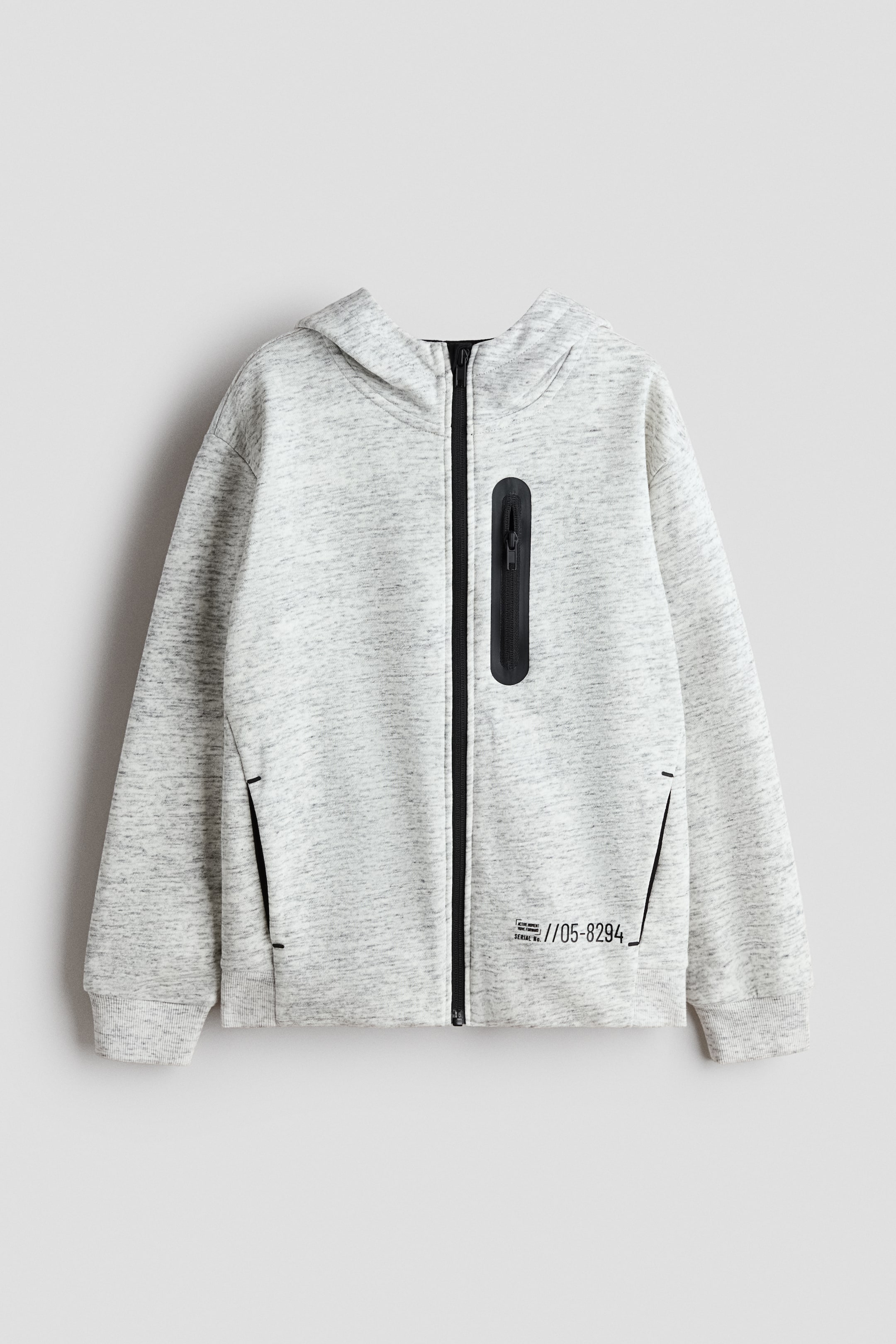 View larger image: A grey marl hooded jacket is shown from the front, featuring a central black zip and a vertical black zip pocket on the wearer’s right chest. Side pockets with black trim are visible, and a small black text graphic appears near the bottom-right hem.