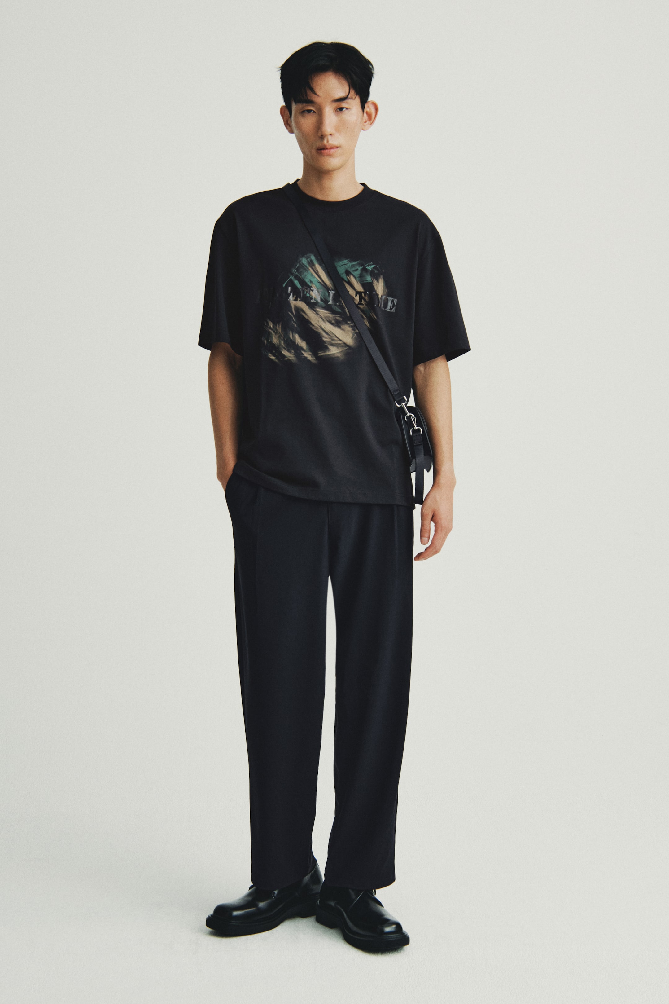 Xem ảnh lớn hơn: Một người đang mặc một chiếc áo phông cổ tròn quá khổ màu đen với họa tiết đồ họa trừu tượng đang đứng đối mặt về phía trước. Nó được phối hợp với quần ống rộng, ôm vừa màu đen, giày đế dày màu đen, và một chiếc túi đeo chéo nhỏ màu đen với phần cứng màu bạc.
