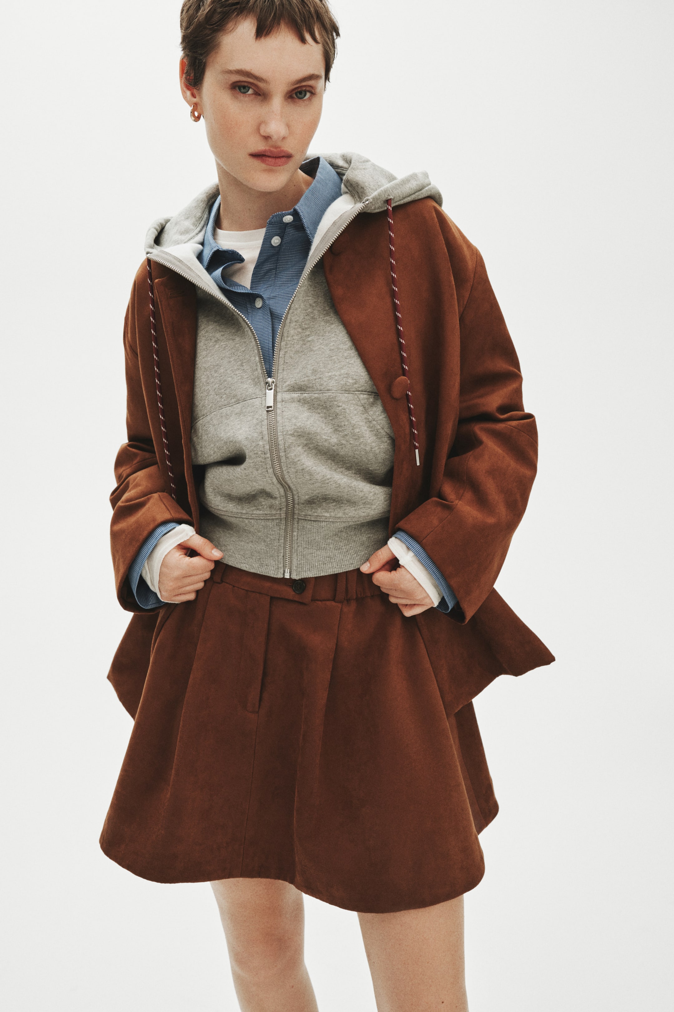 Agrandir l'image: Un blouson marron au toucher doux présente une coupe ample, des manches larges et des cordons décoratifs à l'avant ; il est porté ouvert par‑dessus un sweat à capuche gris zippé. Une jupe assortie taille haute s'évase en de légers plis, complétant l'ensemble coordonné.