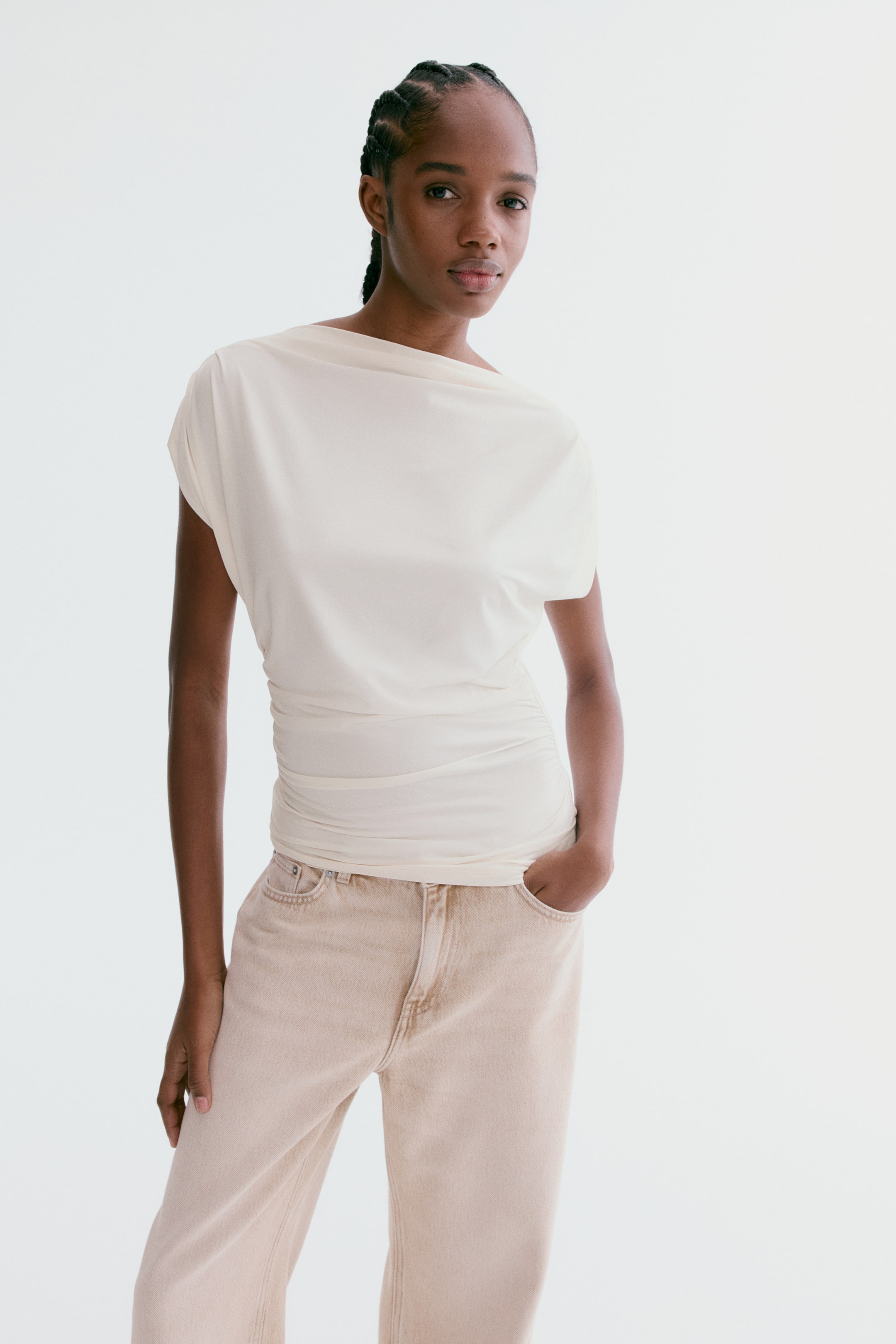 View larger image: The model is wearing an asymmetric jersey top. It has a boat neckline, cap sleeves with pleats on the shoulders, and gathers at the sides of the waist, creating a draped effect. The top is styled with light brown jeans.