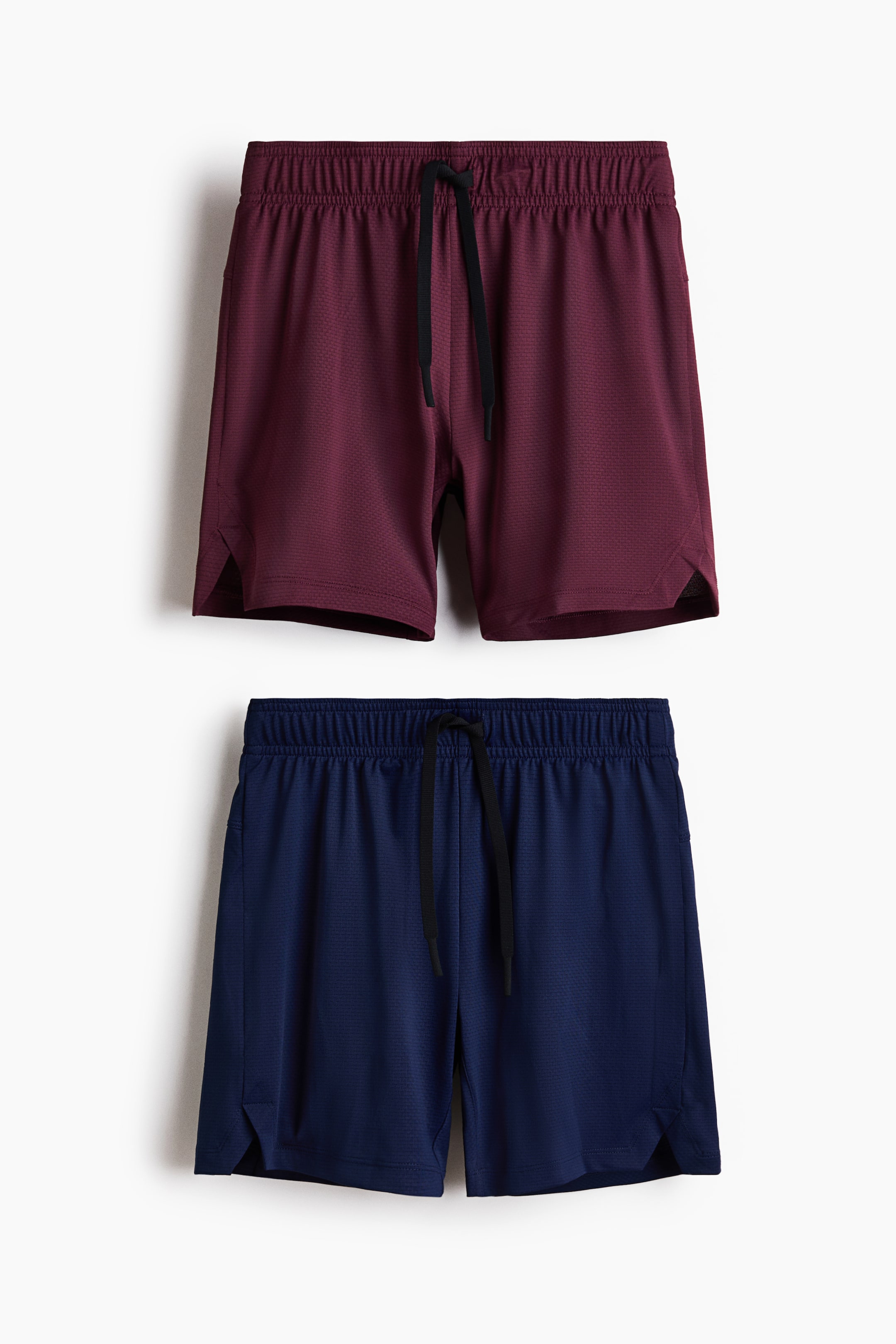 Größeres Bild anzeigen: Zwei Paar Sportshorts sind mit der Vorderseite zu sehen; das obere Paar ist dunkelrot, das untere Paar navyblau. Beide Shorts verfügen über einen elastischen Bund mit schwarzen Kordelzügen, kleinen seitlichen Schlitzen am Saum und bestehen aus einem dezent strukturierten Material.
