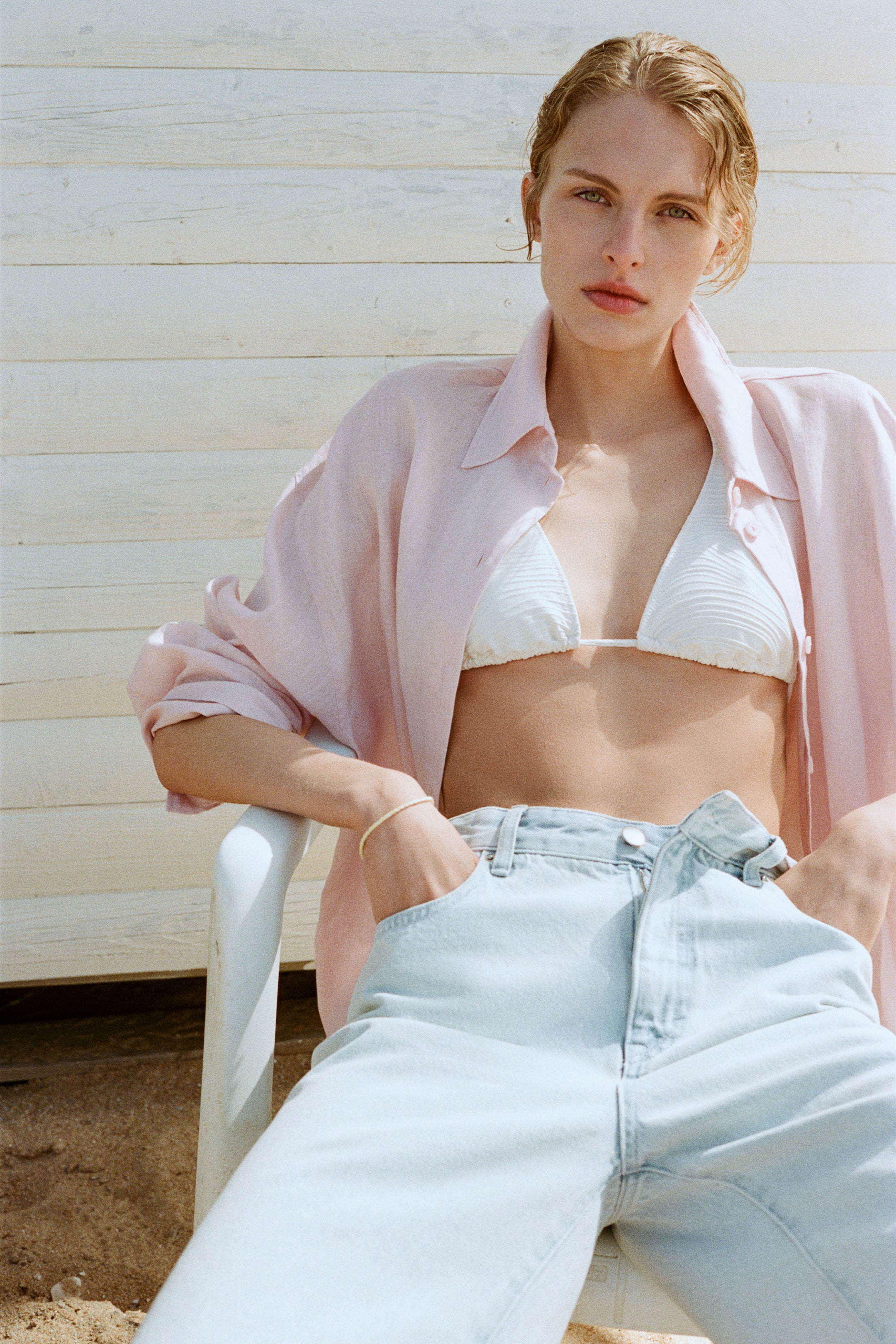 Agrandir l'image: Une chemise col rose clair est portée ouverte et ample par-dessus un haut de bikini triangle blanc côtelé, révélant la taille. Un jean en denim délavé clair, avec une taille haute et des poches avant, est porté avec les mains placées à l'intérieur des poches.