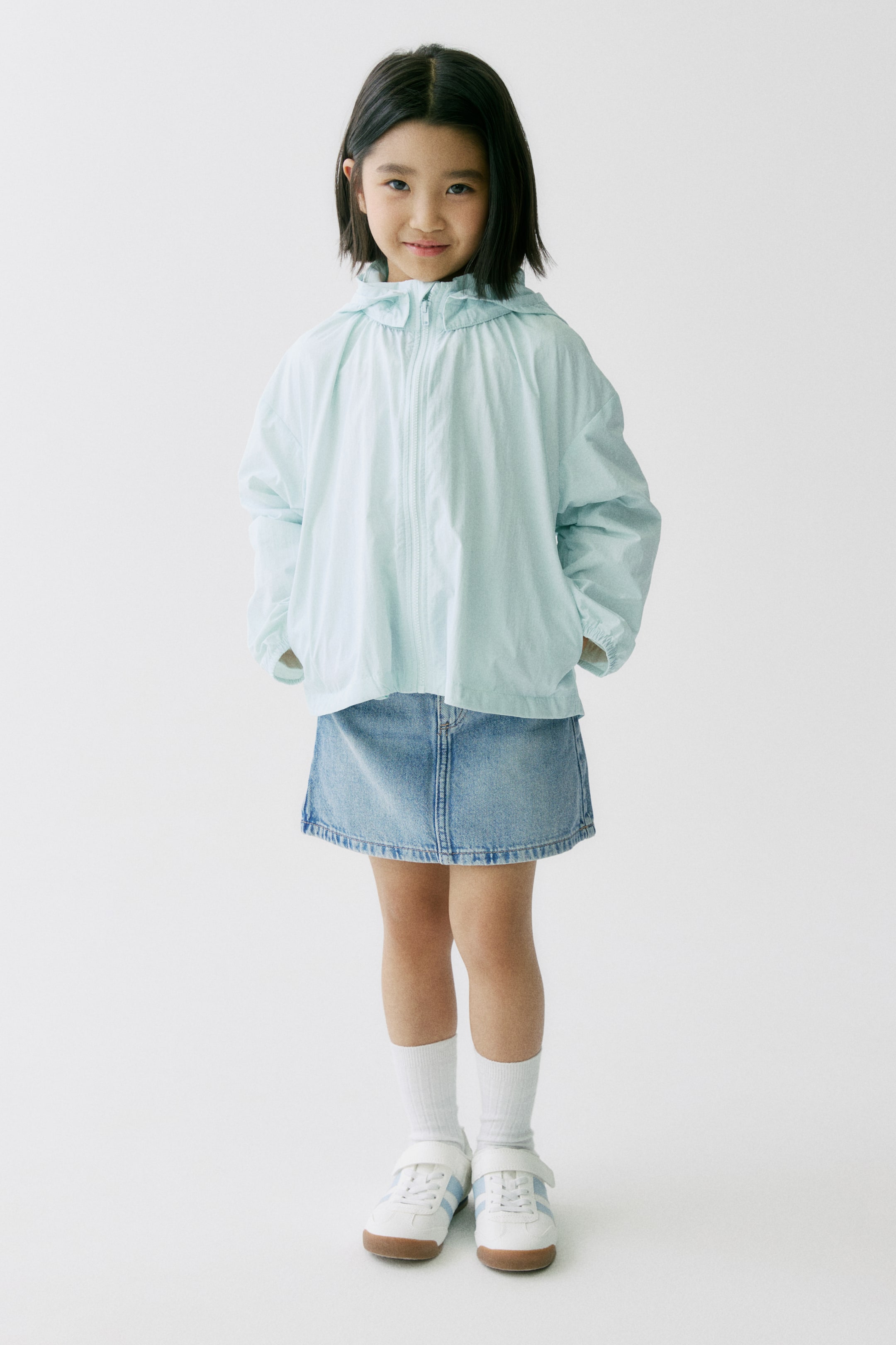 拡大画像を表示: 前から見たライトブルーのフード付きジャケットは、フルジップの前面、胸部に沿った縦のステッチディテール、フード付き、袖口と裾がゴム仕様で、サイドポケットに手を入れた状態です。ライトウォッシュデニムのAラインミニスカートは、ストレートヘムと目立つステッチが特徴です。