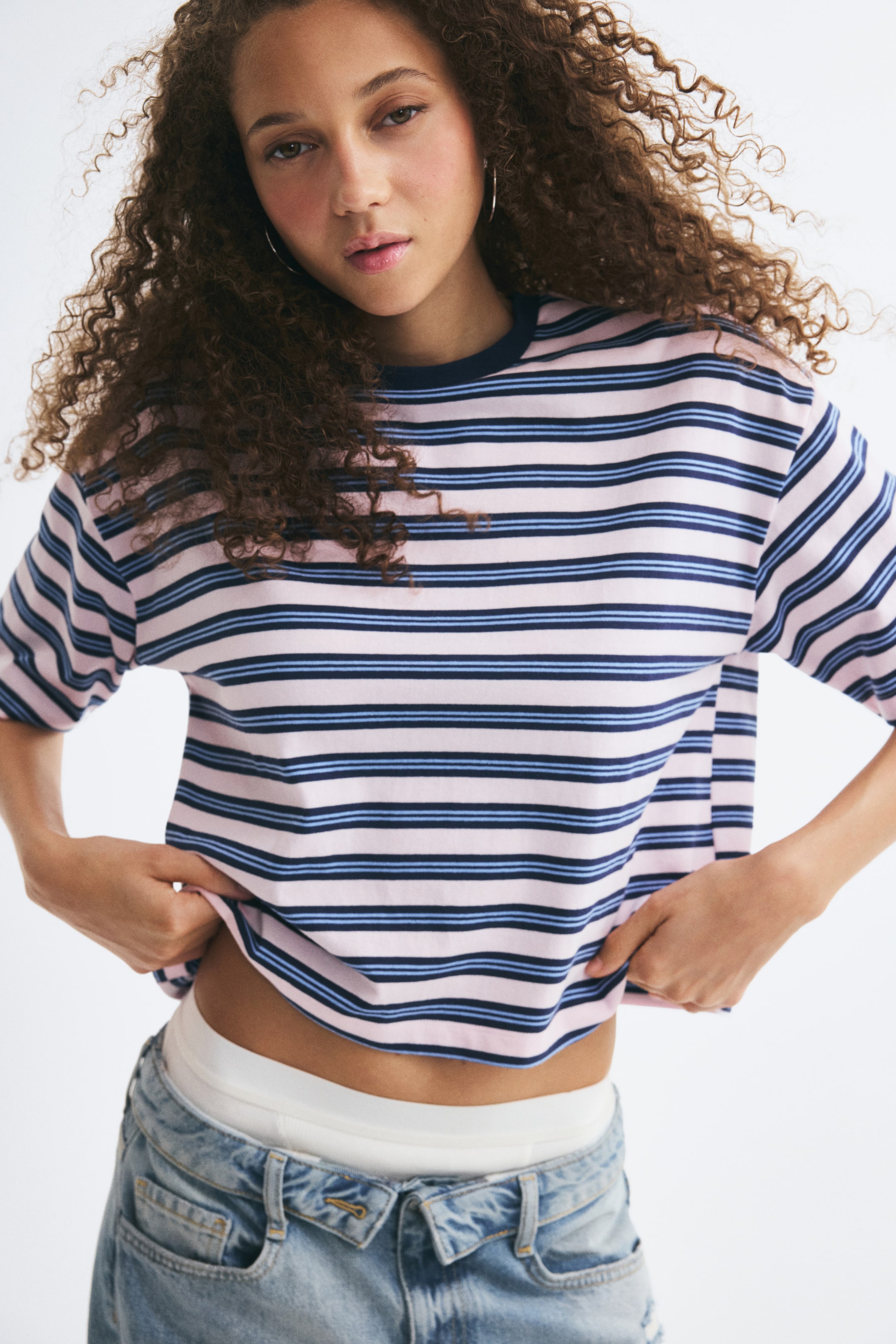 View larger image: An oversized T-shirt with pink, light blue, and navy horizontal stripes is worn by the model, who is holding up the hem with both hands. The T-shirt features a navy blue rib-trimmed crew neck, dropped shoulders, and elbow-length sleeves, and is styled with light-wash jeans.