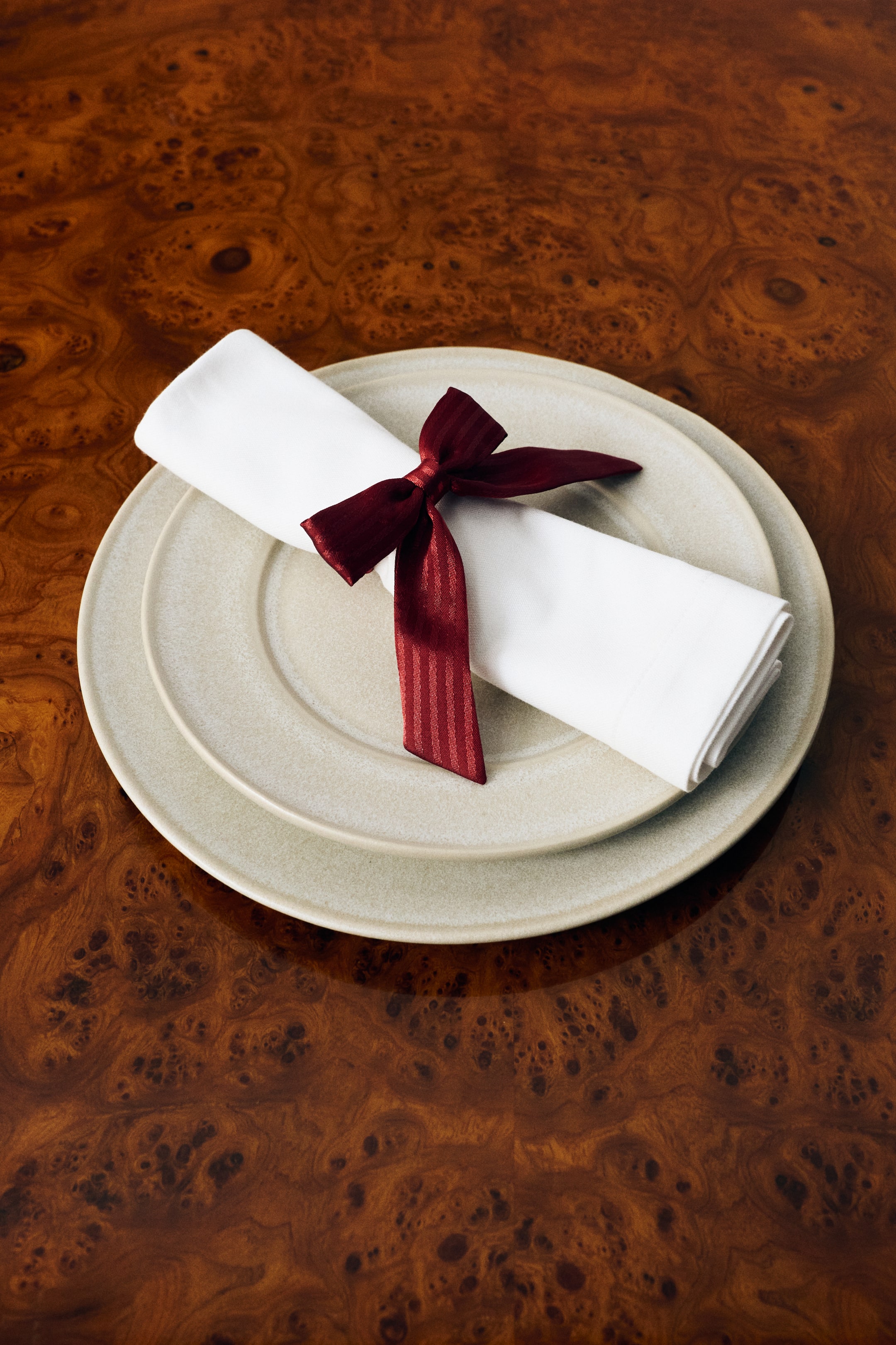 View larger image: A burgundy napkin ring covered in a shiny weave and featuring a bow detail at the front is holding a folded white napkin. The napkin and ring are resting on a set of stacked light beige ceramic plates.