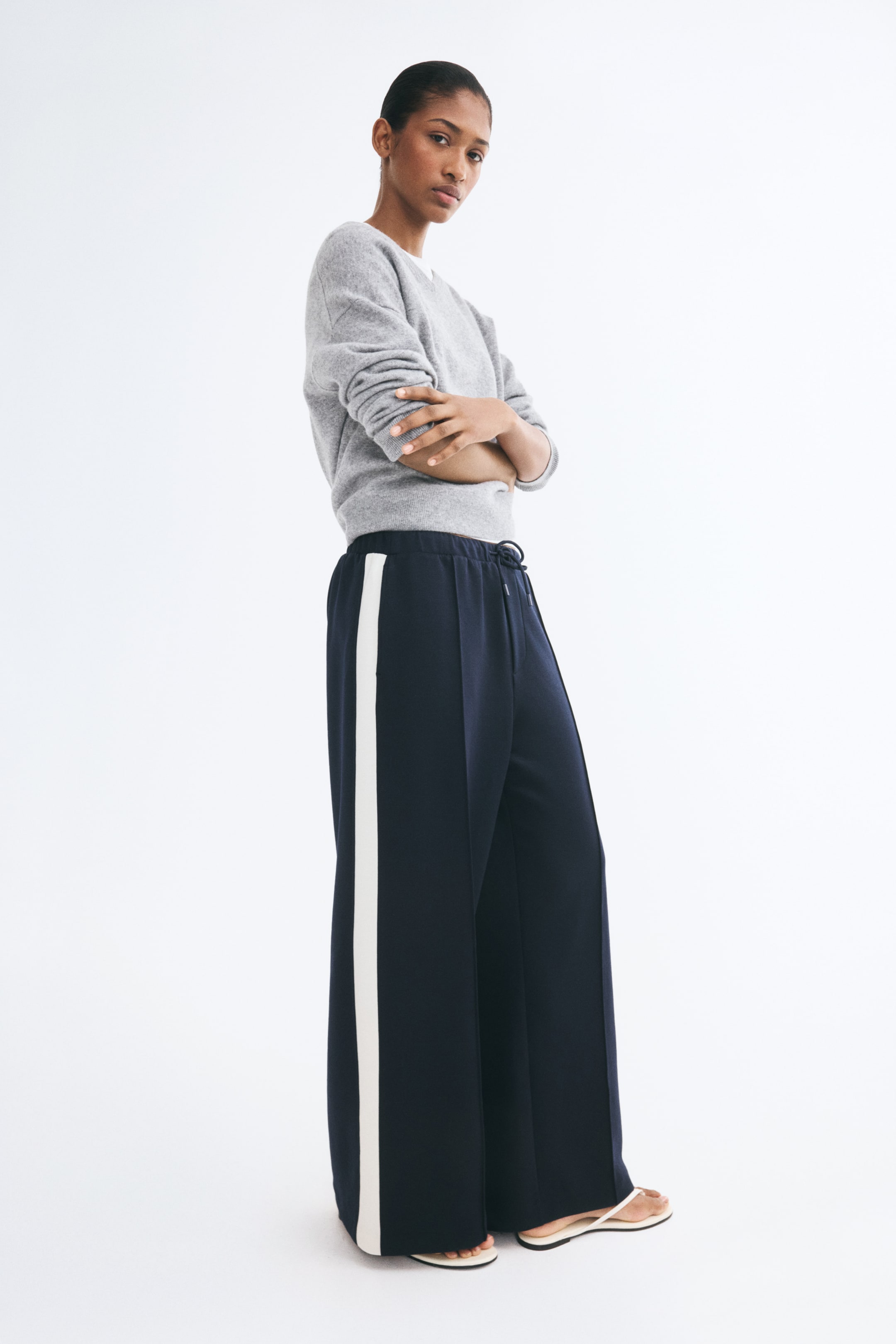 Ampliar la imagen: La modelo lleva unos pantalones tipo jersey de pierna ancha en color azul marino, con una franja blanca contrastante a lo largo del costado, cintura elástica con cordón ajustable y pliegues frontales cosidos. Los pantalones se combinan con un suéter gris de cuello redondo y sandalias planas.