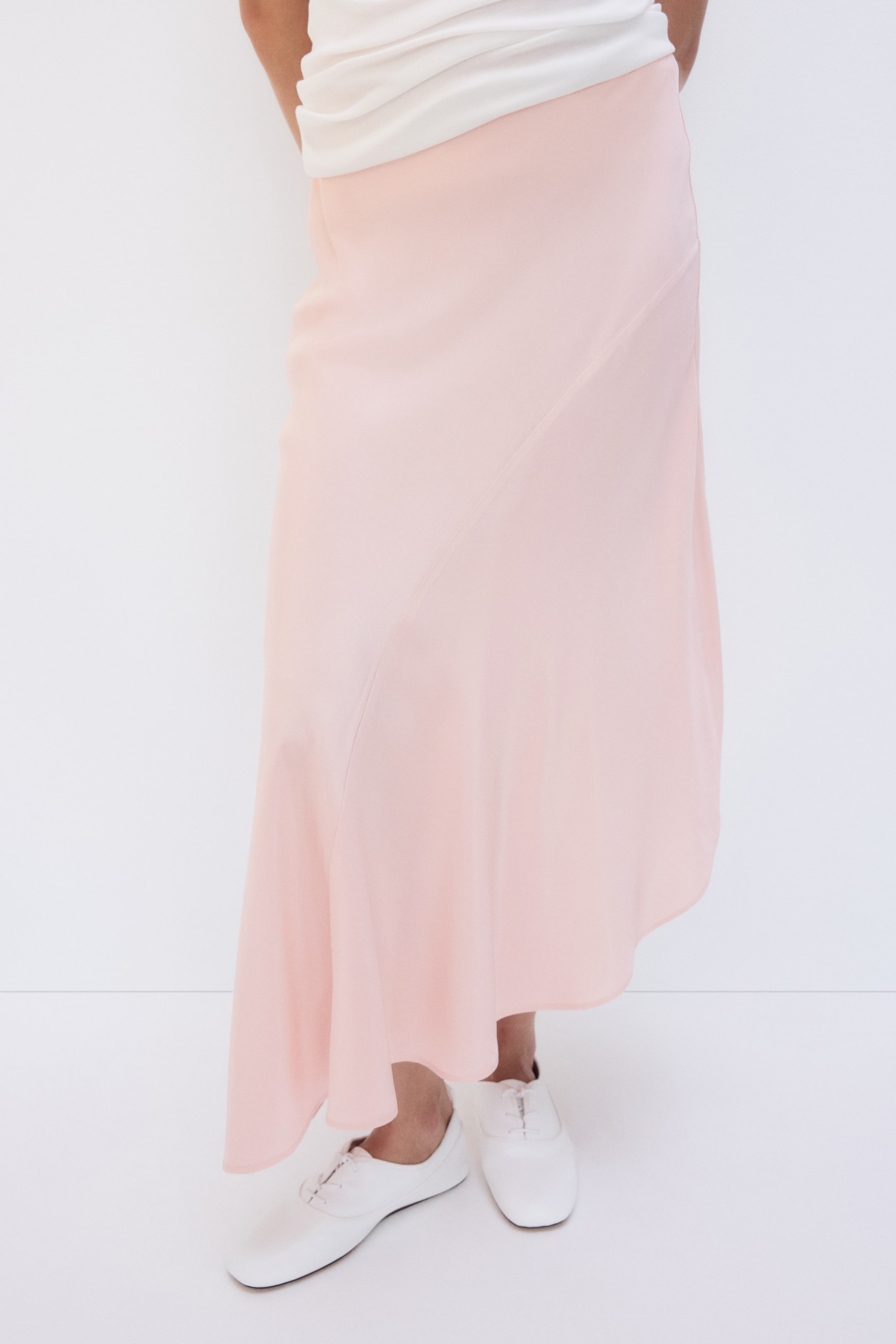 Ampliar la imagen: La modelo lleva una falda de satén rosa claro, de largo a la pantorrilla, que destaca por su tela de caída suave, costura diagonal curva y dobladillo asimétrico. La falda se combina con una blusa blanca drapeada y zapatos blancos con agujetas.