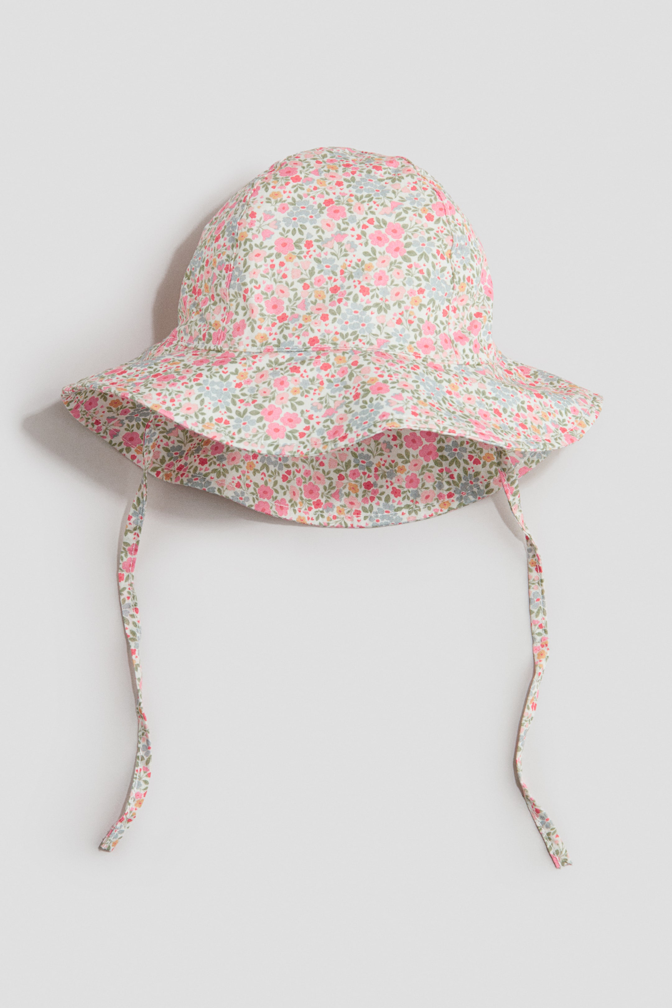 Ver imagen más grande: Un sombrero de sol se presenta de frente, mostrando un estampado floral colorido con flores rosas, azul claro y amarillas sobre un fondo claro. Está diseñado con un ala suave e inclinada, y dos lazos de tela a juego cuelgan por debajo del ala.