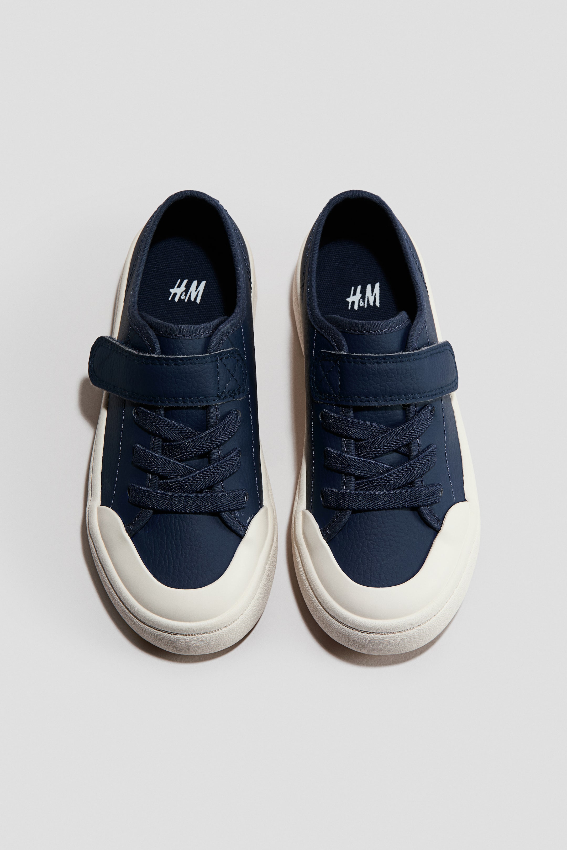 Agrandir l'image: Une paire de chaussures bleu marine est présentée de face, chacune avec une tige texturée, un embout en caoutchouc blanc cassé proéminent et des semelles épaisses. Une sangle velcro bleu marine sécurise chaque chaussure par-dessus des lacets factices, et le logo H&M est visible sur les semelles intérieures bleu marine.