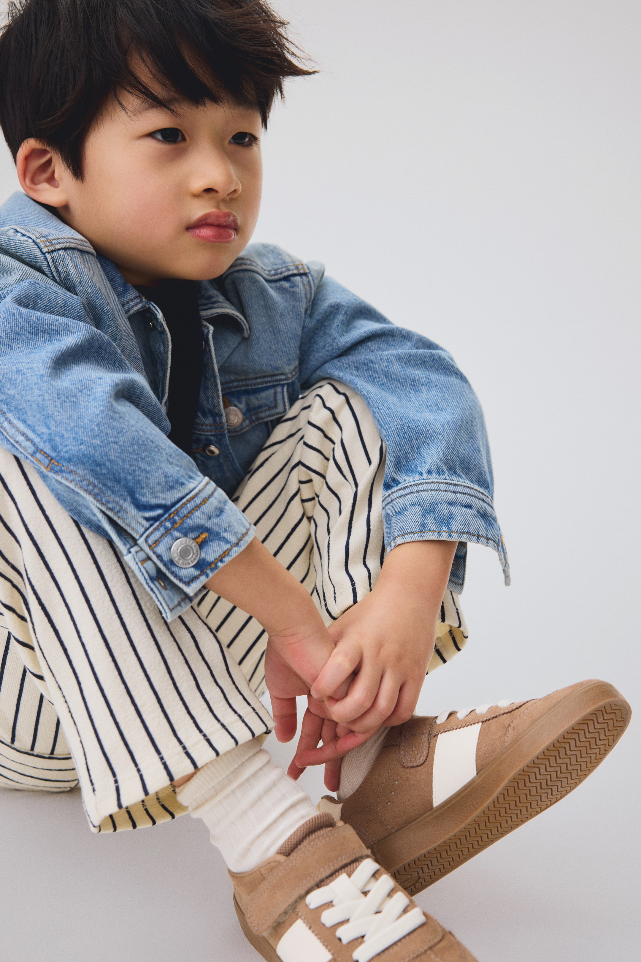 Größeres Bild anzeigen: Eine hell gewaschene Jeansjacke mit Metallknöpfen wird getragen, über einem schwarzen T-Shirt geschichtet. Cremefarbene Hosen mit vertikalen dunklen Streifen werden getragen, und beige Sneakers mit weißen Seitenstreifen und weißen Schnürsenkeln werden getragen, seitlich abgewinkelt, wodurch cremefarbene gerippte Socken sichtbar werden.