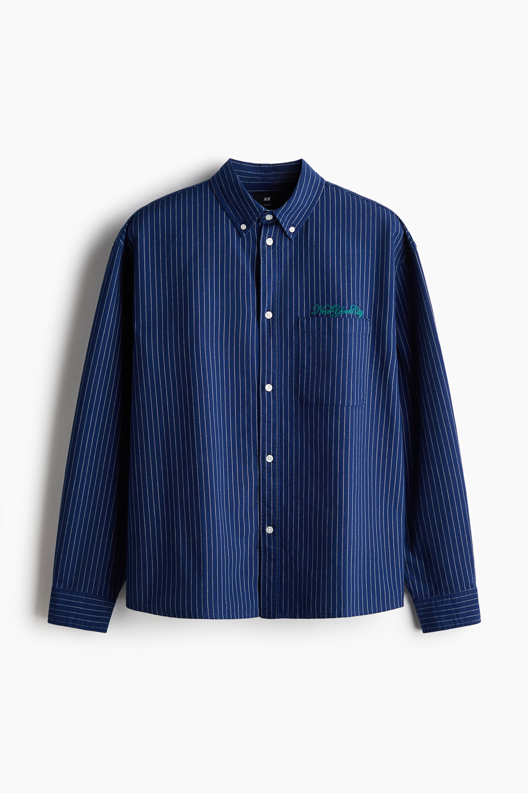 View larger image: A navy blue pinstriped Oxford cotton shirt is shown from the front, featuring a button-down collar, a classic buttoned front, and an open chest pocket on the left side with a green 'New York City' embroidered motif above it. Long sleeves end with buttoned cuffs and a sleeve placket, while a straight-cut hem completes the relaxed fit.