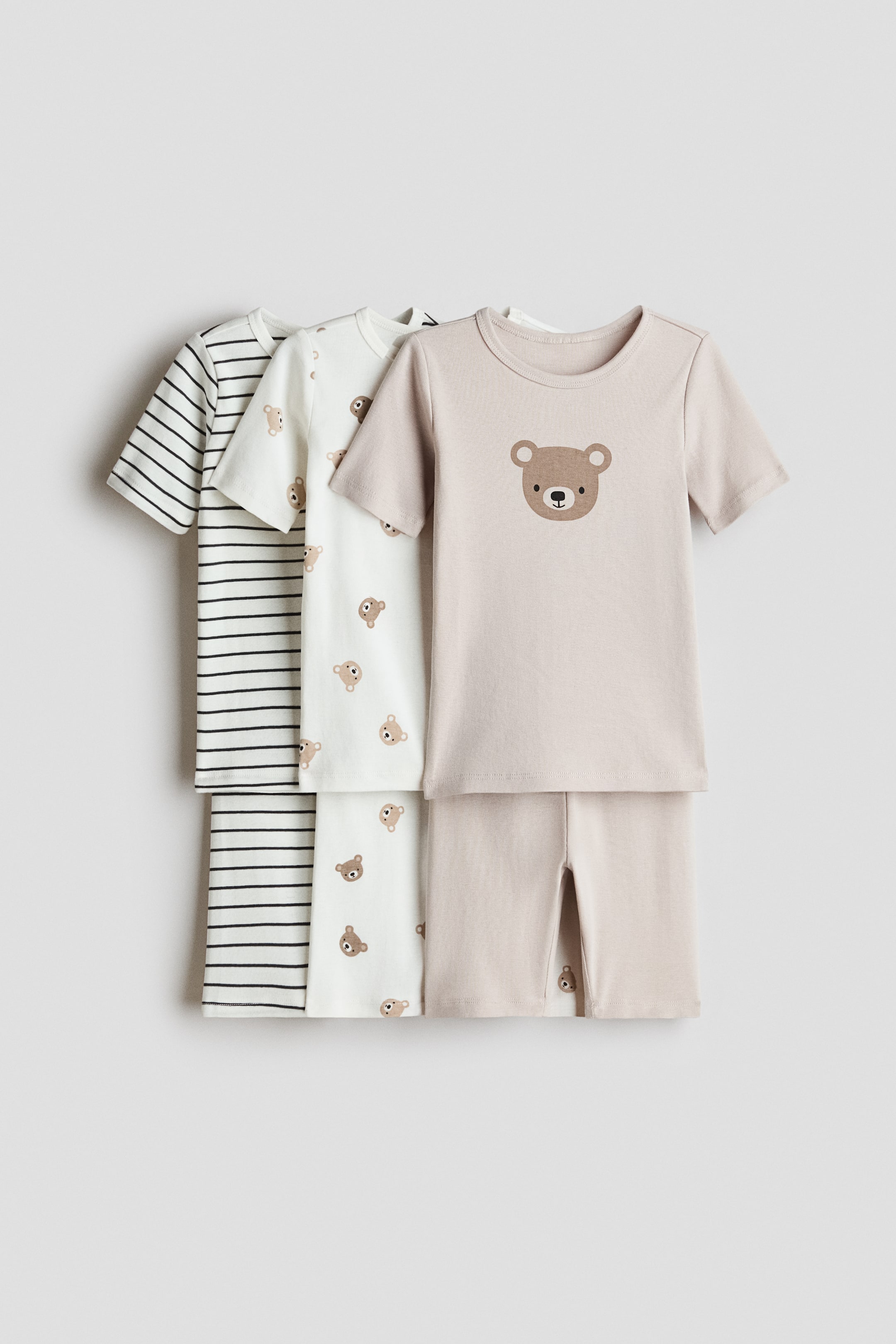 View larger image: Three sets of baby pajamas are displayed in layers, each set comprising a short-sleeved top and matching long pants. The front set is beige, featuring a large bear face graphic on the top and a small bear face on the bottom's inner leg. The middle set showcases an off-white fabric with an all-over small bear face pattern, and the back set displays an off-white fabric with horizontal black stripes.
