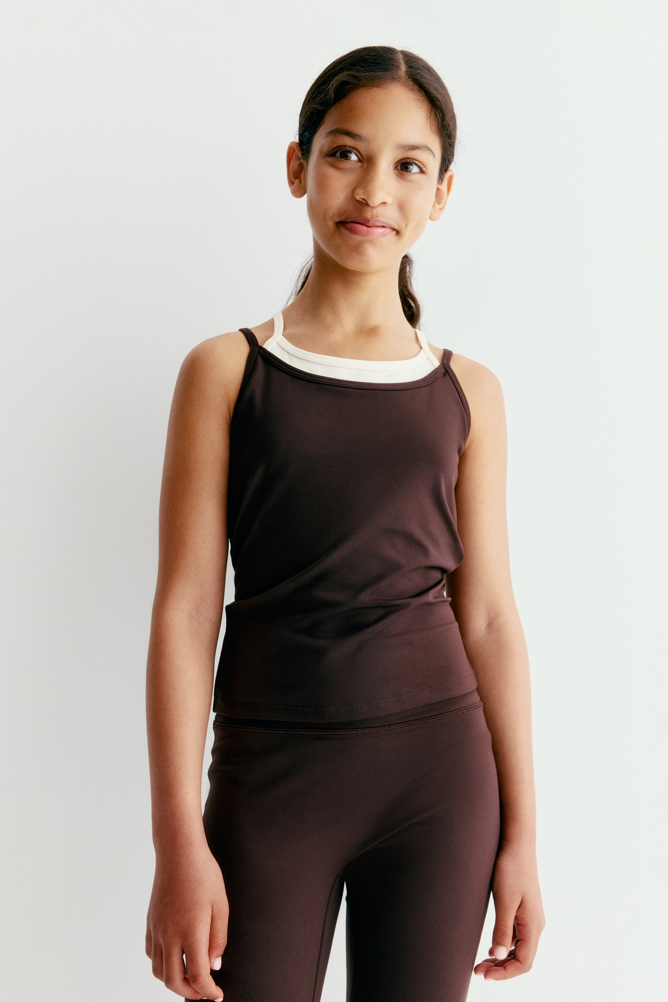 Größeres Bild ansehen: Ein dunkelbraunes Athletic-Top mit dünnen Trägern ist von vorne zu sehen und verfügt über eine figurbetonte Silhouette sowie einen Lagenlook über einem cremefarbenen Tanktop. Das Oberteil wird mit einer passenden dunkelbraunen Leggings kombiniert und bildet so ein abgestimmtes Activewear-Set.