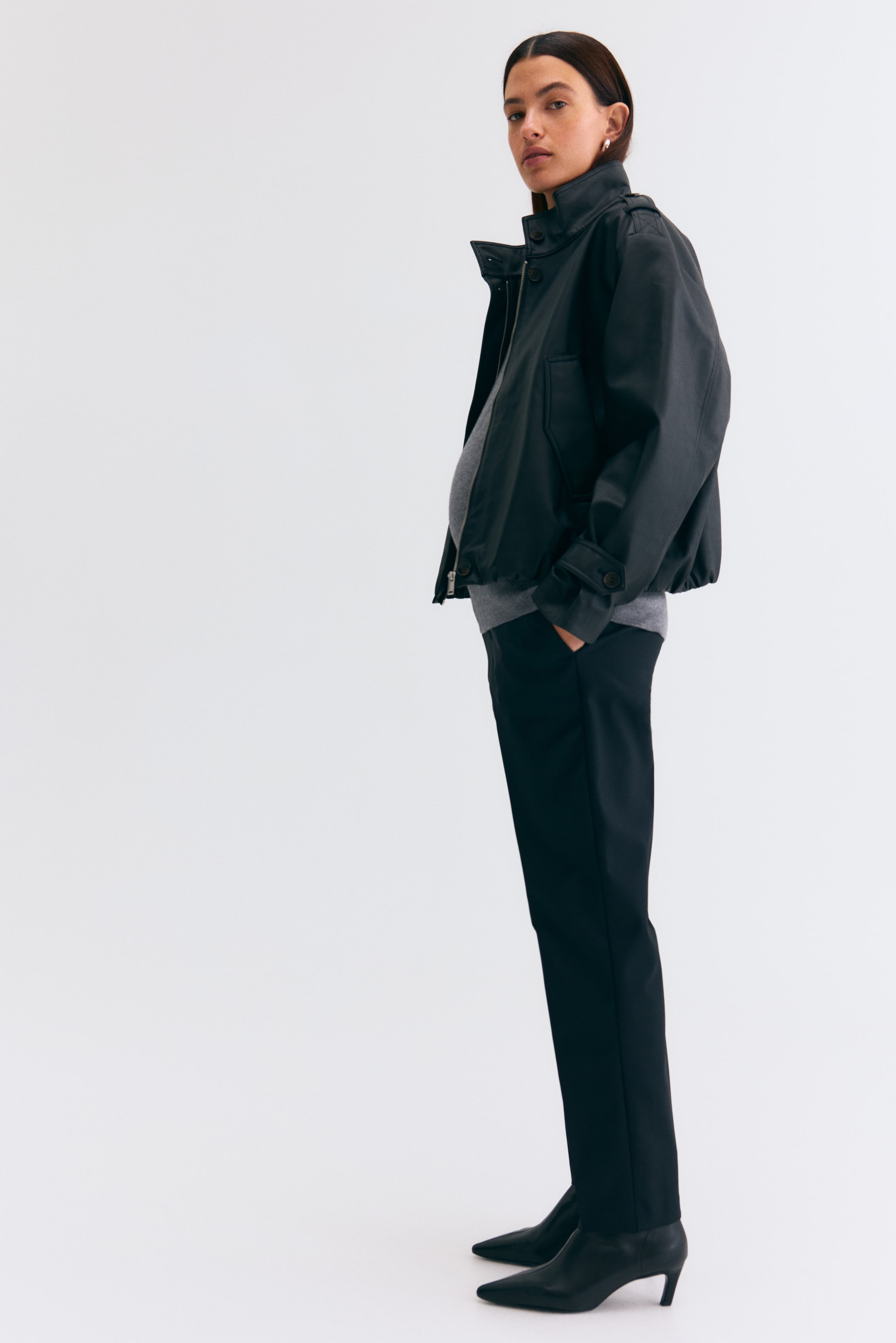 Agrandir l'image: Une veste bomber noire est présentée de profil, orientée vers la droite, avec un col montant fermé par des pressions, une fermeture éclair frontale et des manches longues munies de poignets à pressions réglables. La veste comprend également des poches avant à rabat, un ourlet élastiqué et est portée par-dessus un haut gris visible.